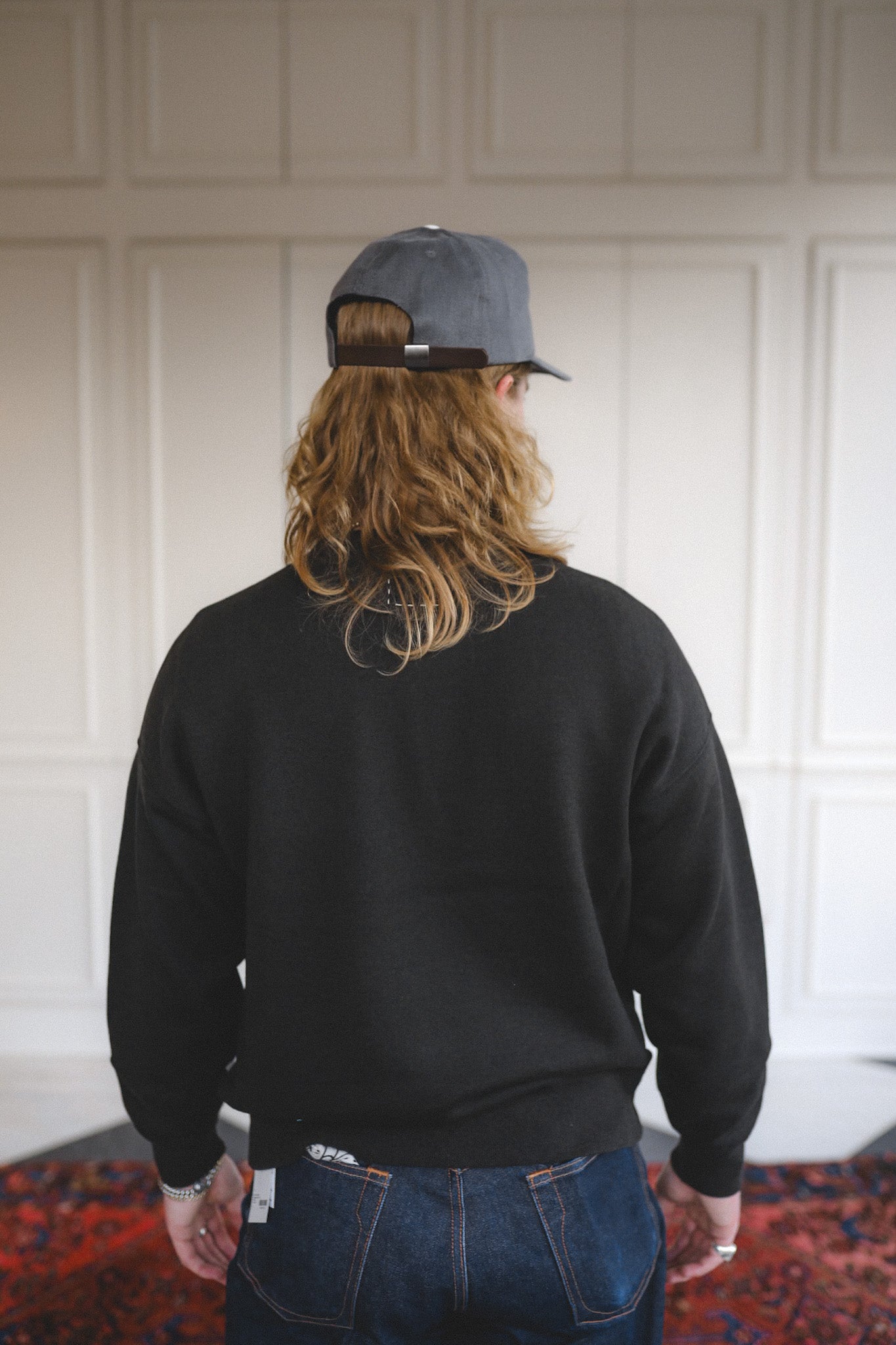 Person wearing a black sweatshirt and blue jeans with a cap, standing in a room with white paneled walls.