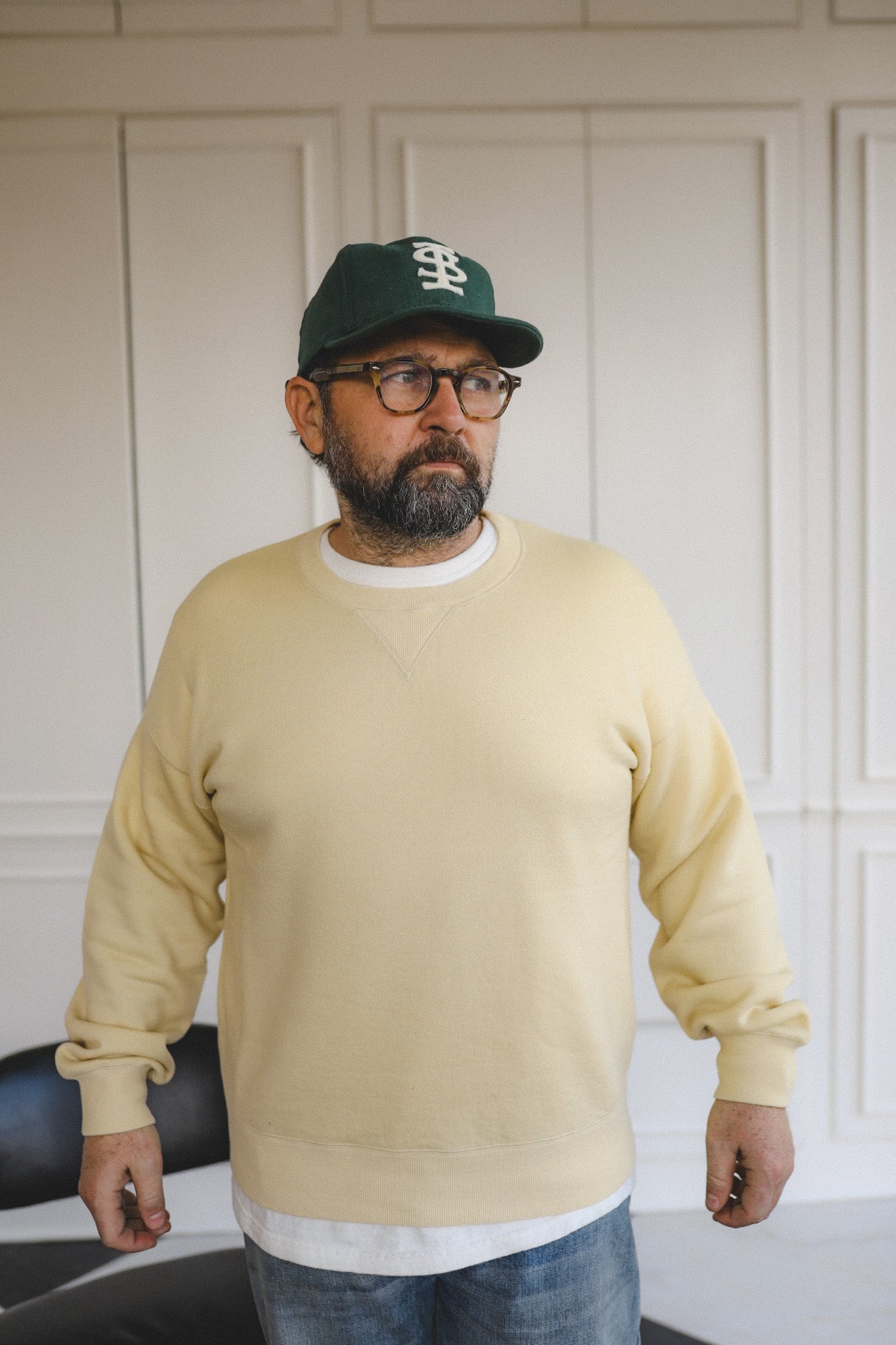 Man wearing a beige sweatshirt, dark cap, and glasses against a white paneled wall.