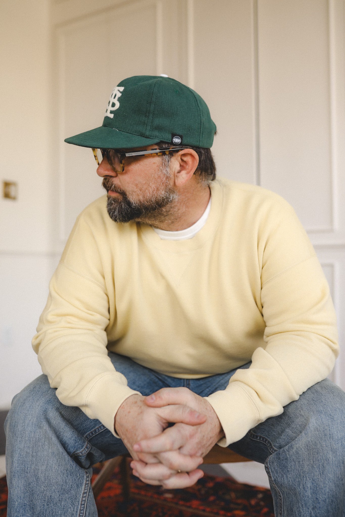 Man wearing a green cap and beige sweater sitting on a chair.