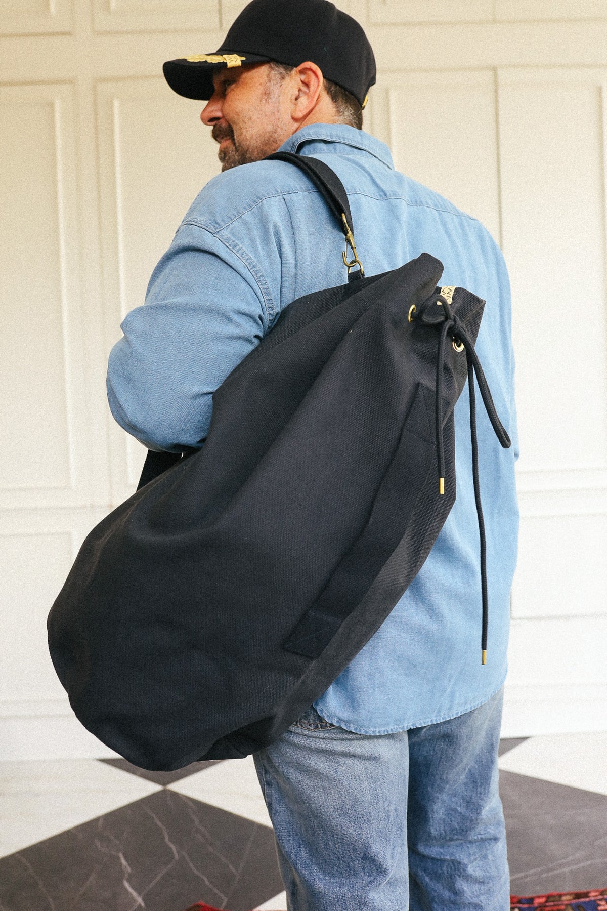 A man wearing a light blue denim shirt and jeans, seen from behind, carrying a large black canvas drawstring bag over his shoulder.