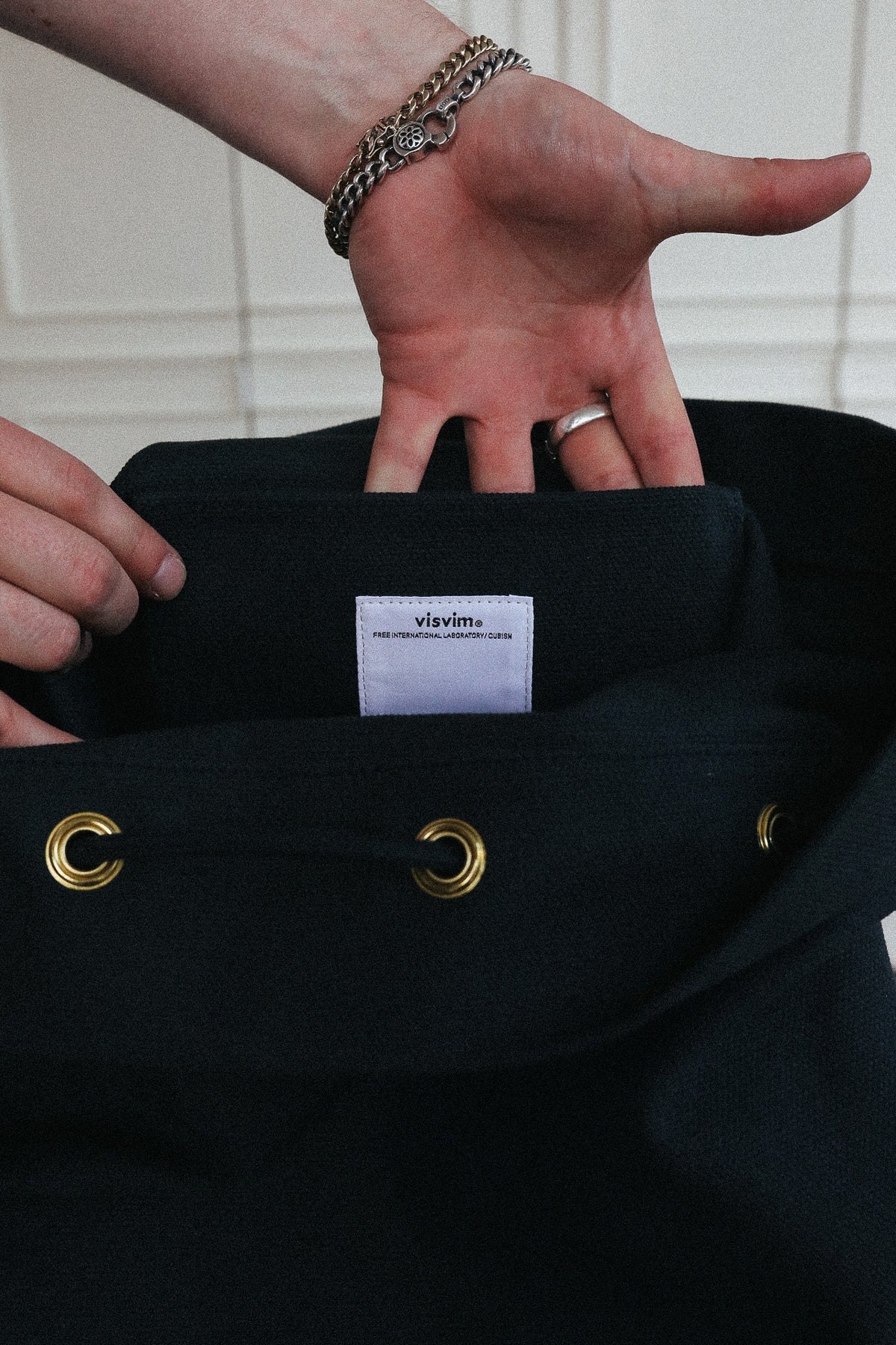 A close-up of a person's hands holding open a black drawstring bag, showing a white brand label that reads 'vixén'. The bag has gold-tone metal grommets and is photographed against a light tiled background.