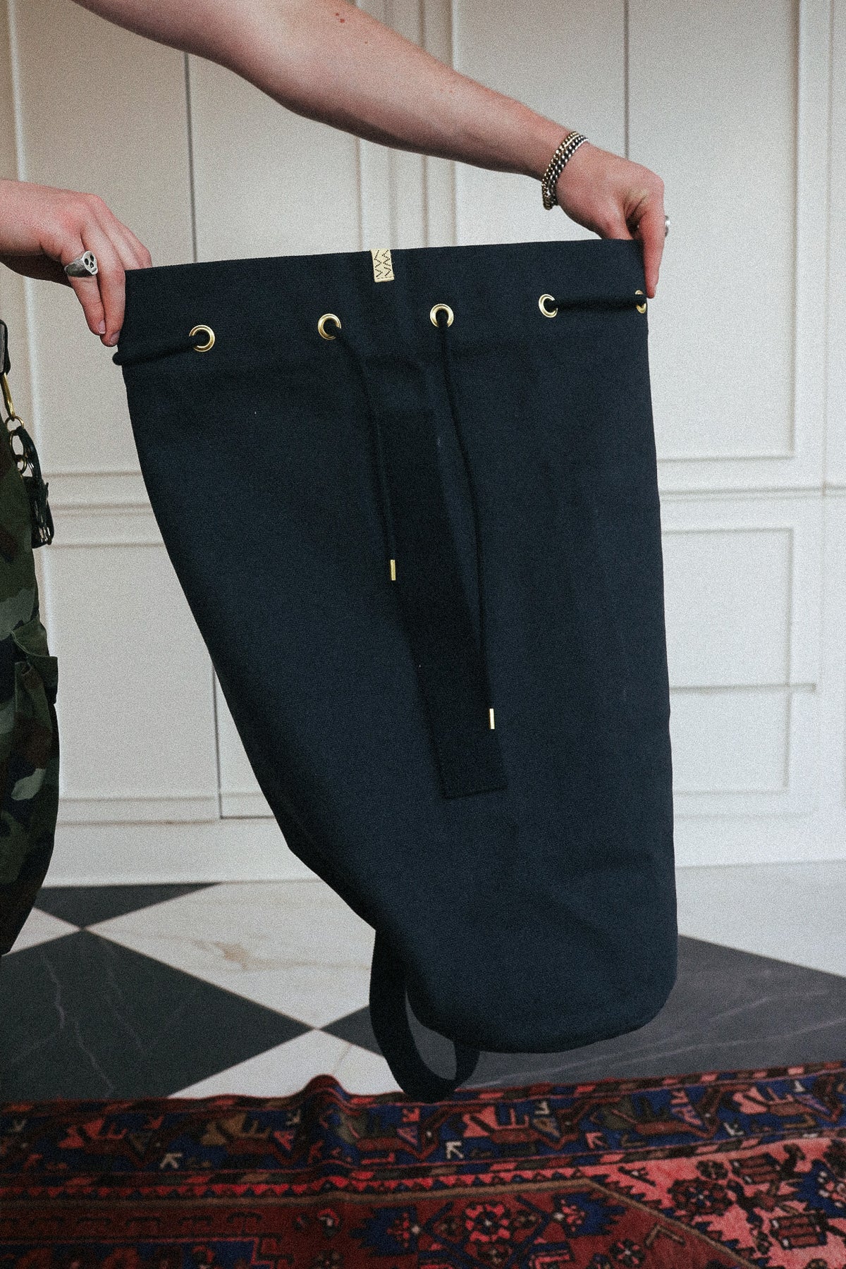 A person holds up a large black cylindrical bag or container with gold-tone grommets and a hanging strap, photographed indoors against a white paneled wall and patterned rug.