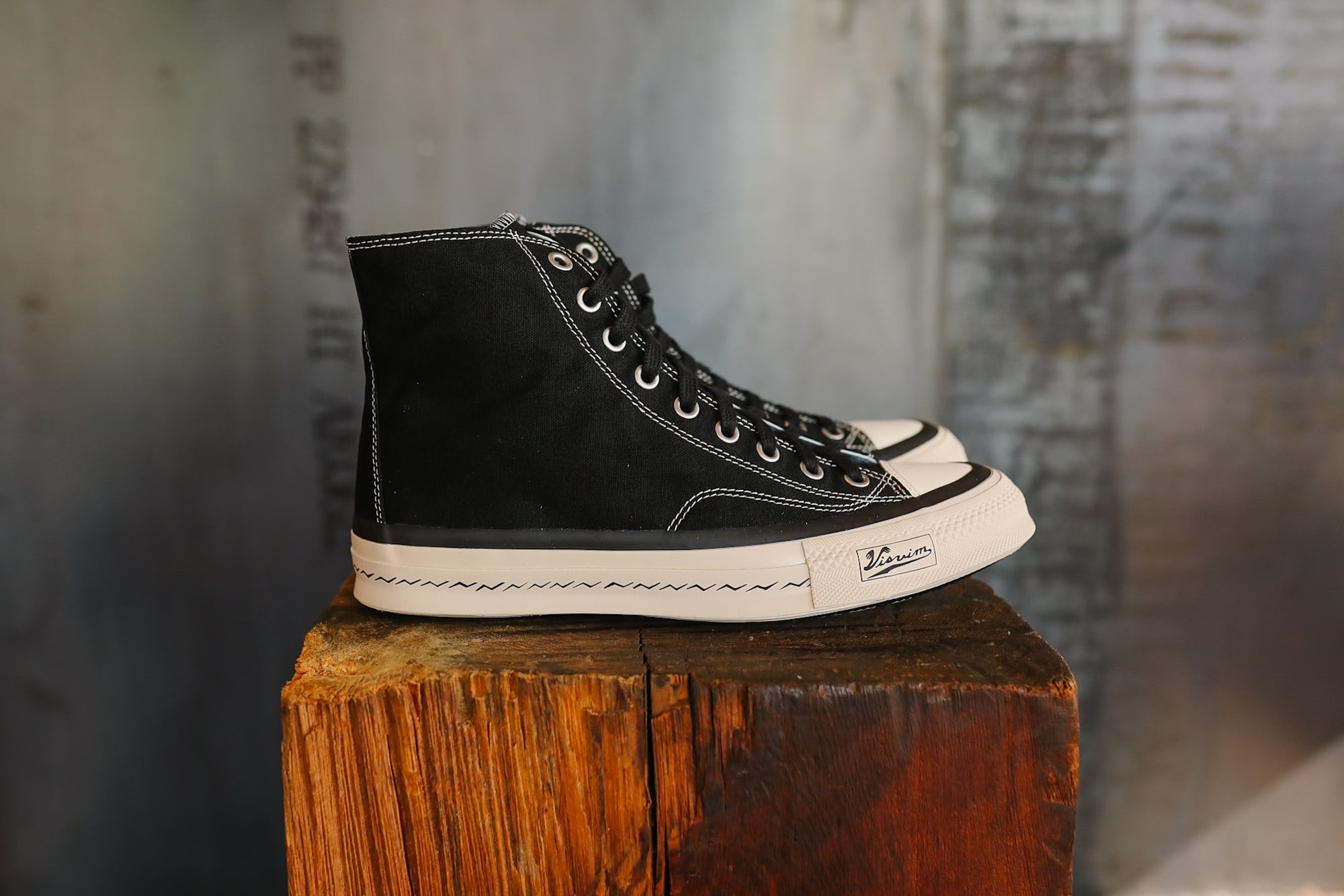 Black high-top sneaker with white sole on a wooden block against a textured wall.