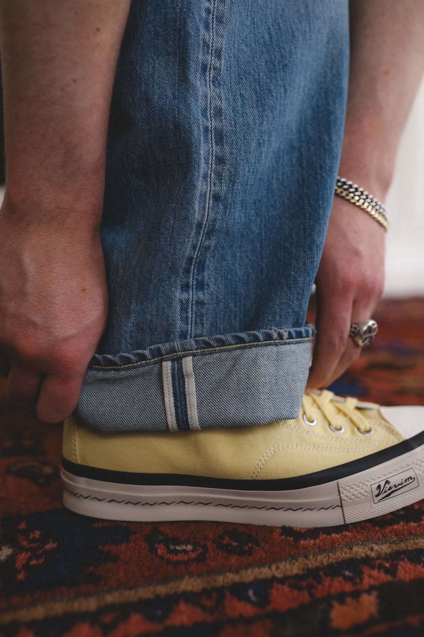 Person wearing yellow sneakers with rolled-up blue jeans on a patterned rug.