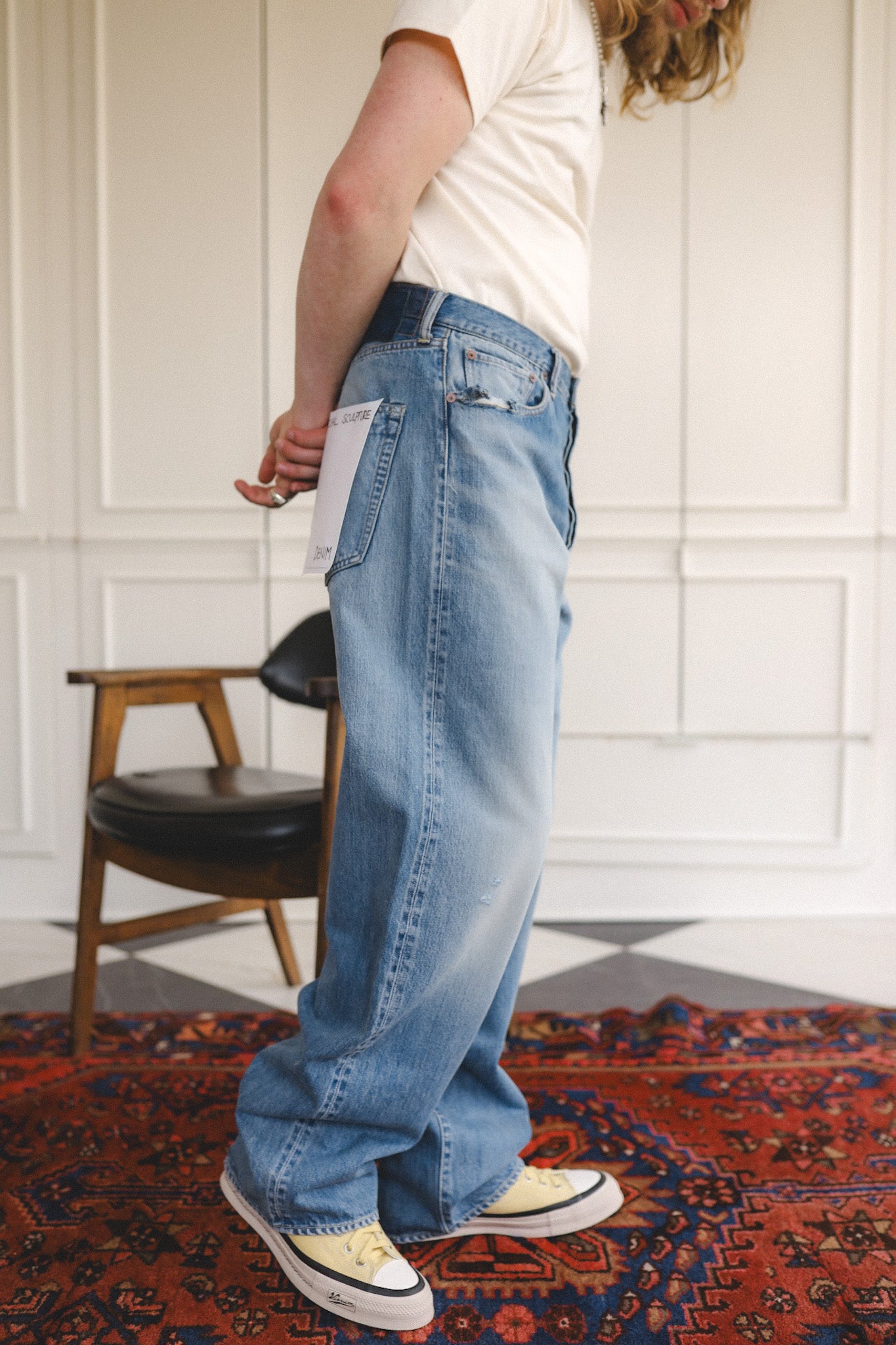 Person wearing blue jeans and a white shirt standing on a patterned rug with a chair in the background.
