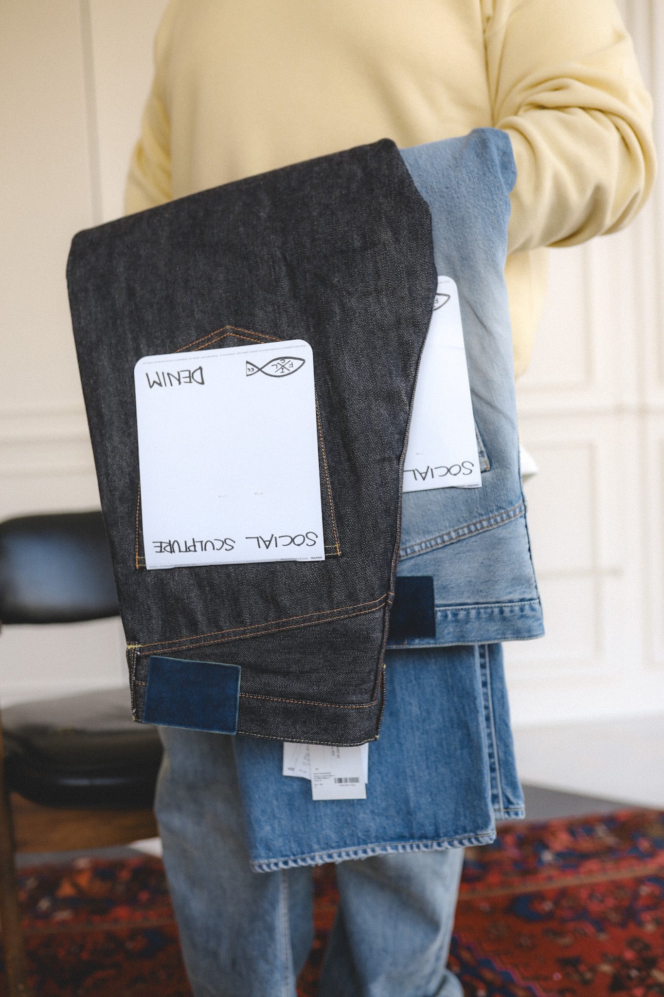 Person holding two pairs of jeans with labels on a patterned rug.