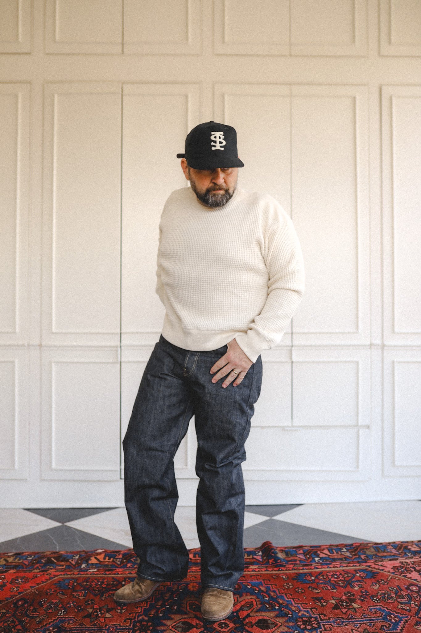 Man wearing a white sweater and dark jeans standing in a room with a patterned rug and white paneled walls.