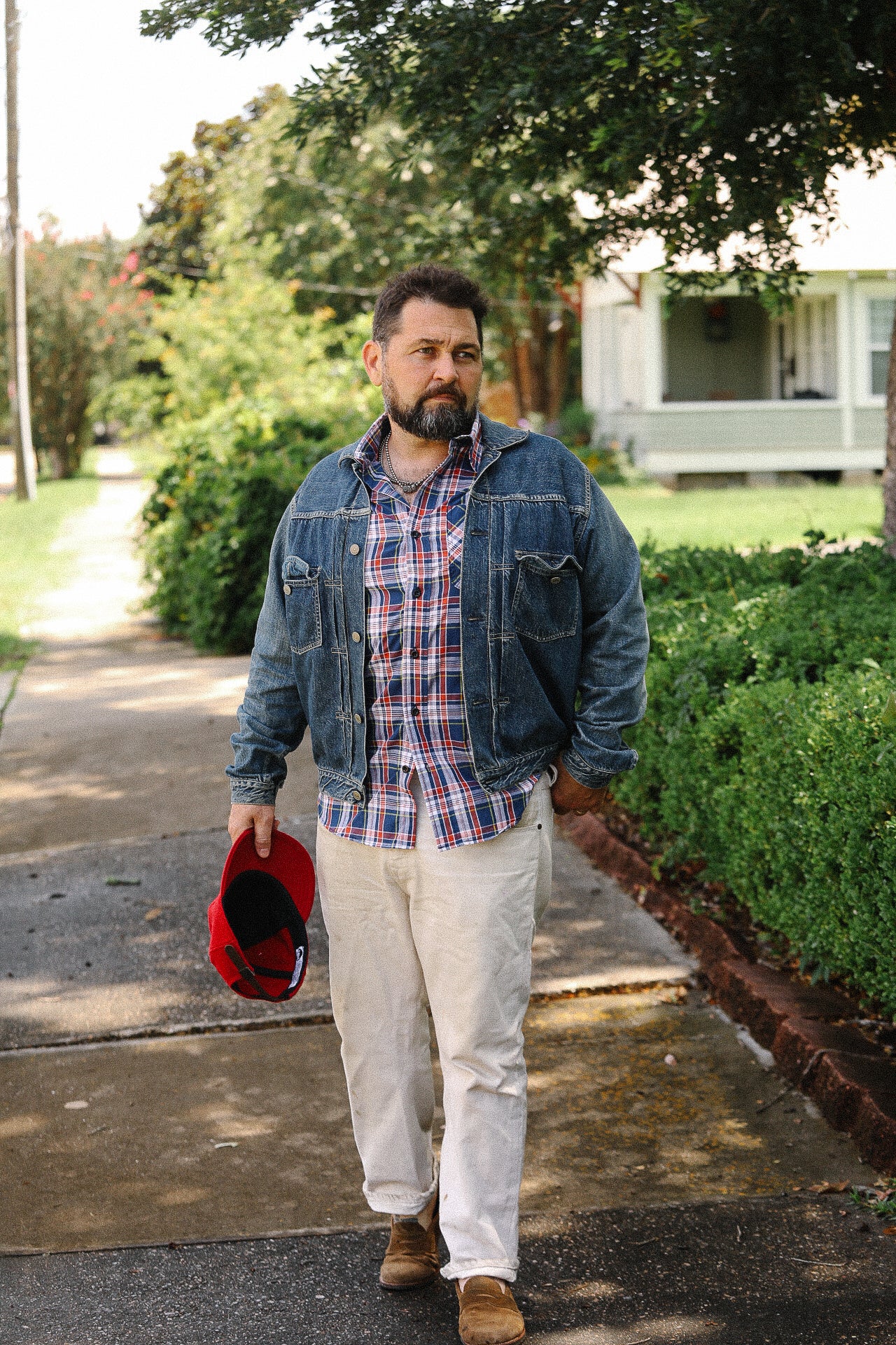 Man wearing denim jacket, plaid shirt, beige pants, holding red cap, walking on sidewalk in a residential neighborhood