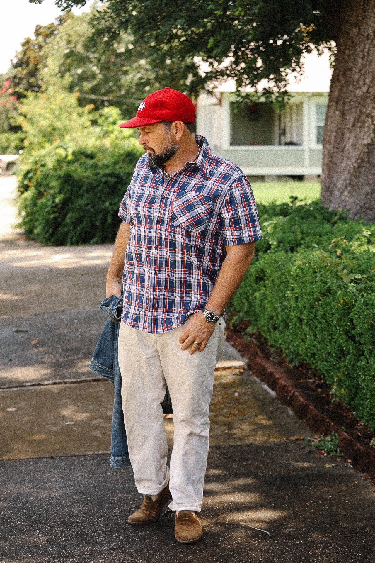Man wearing navy red plaid short sleeve madras shirt, beige pants, brown shoes, and red cap outdoors