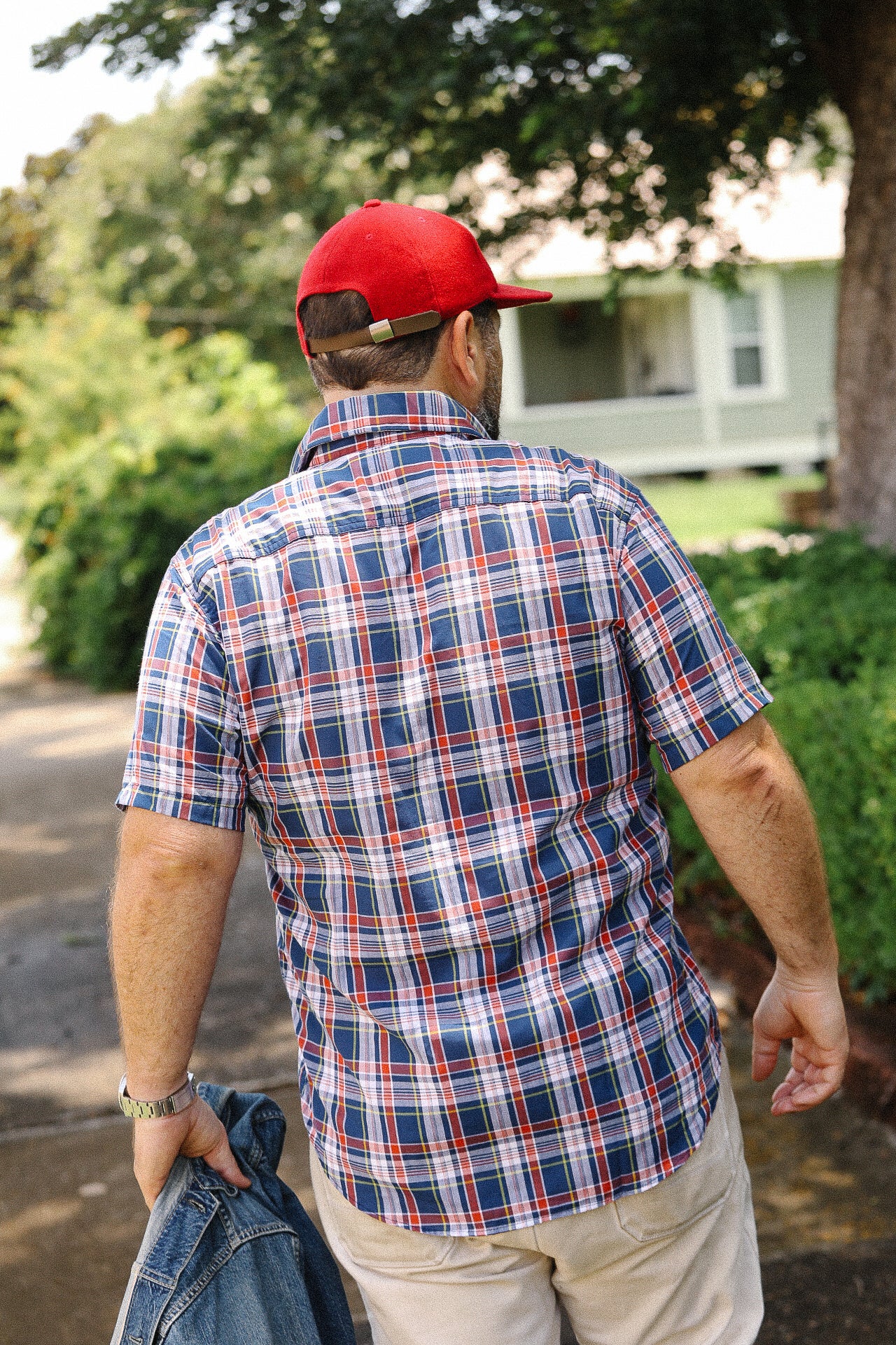 Man wearing navy red short sleeve madras plaid shirt and red baseball cap walking outdoors with greenery