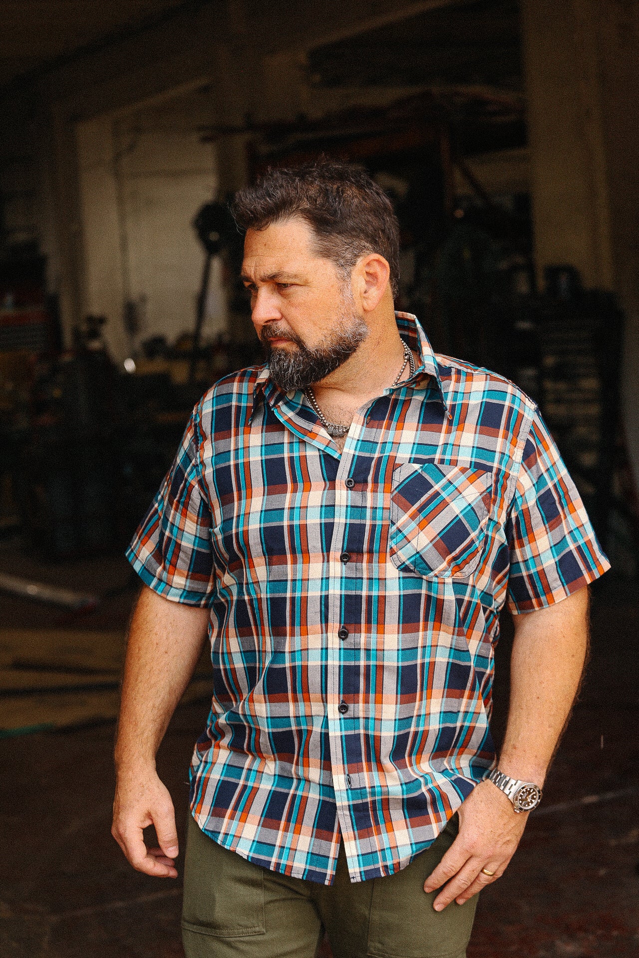 Bearded man wearing turquoise, orange, and navy plaid short sleeve shirt in rustic workshop setting