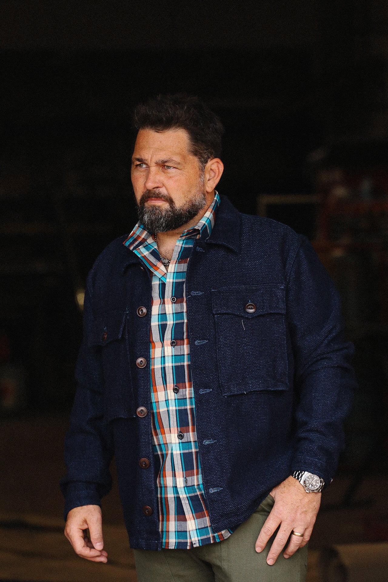 Bearded man wearing turquoise and orange plaid shirt under dark blue buttoned jacket with olive pants in workshop