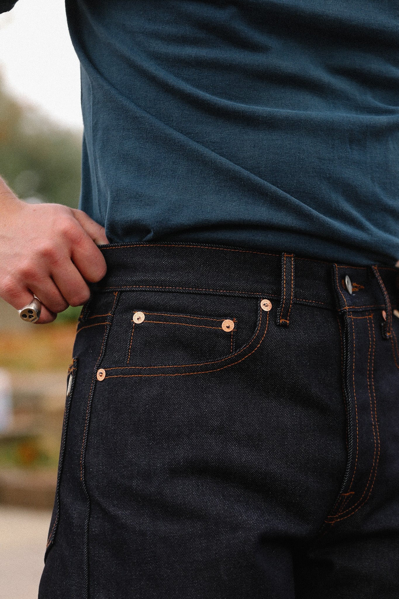 Close-up of a person wearing dark indigo raw denim jeans with contrast orange stitching and a teal shirt