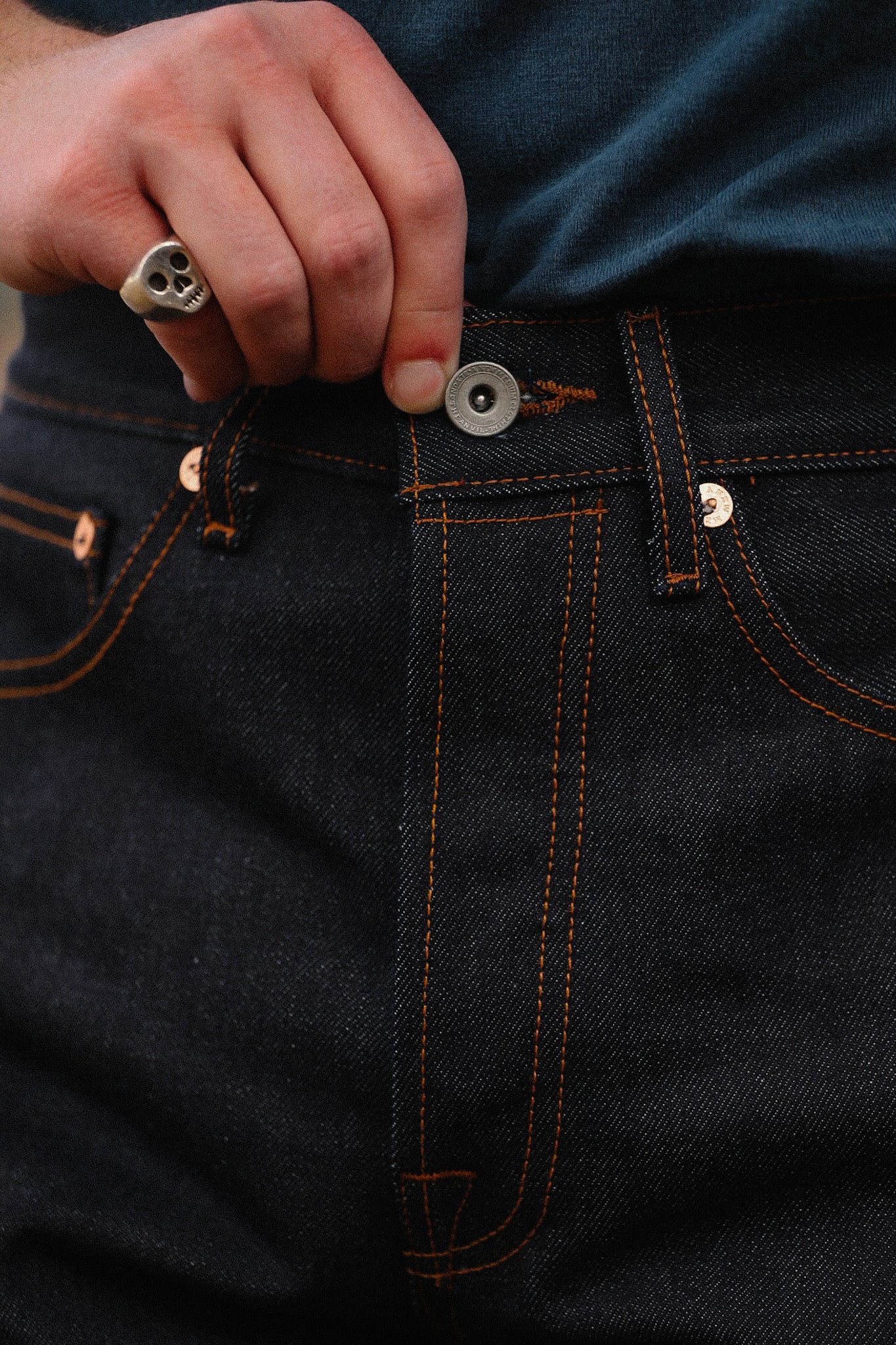 Close-up of hand with skull ring fastening button on dark indigo raw denim jeans with orange stitching