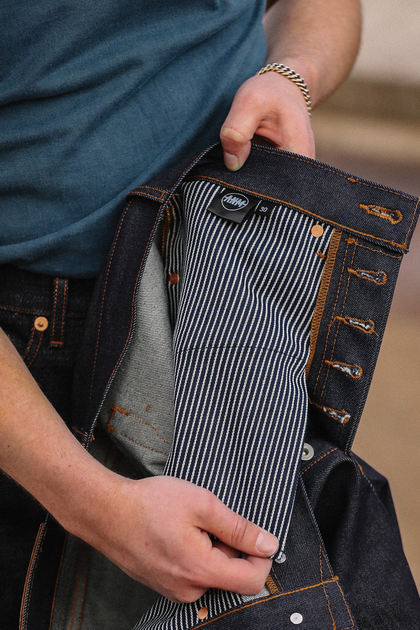 Close-up of hands holding Indigo 16.5oz Kaisen denim jeans showing striped inner lining and button fly