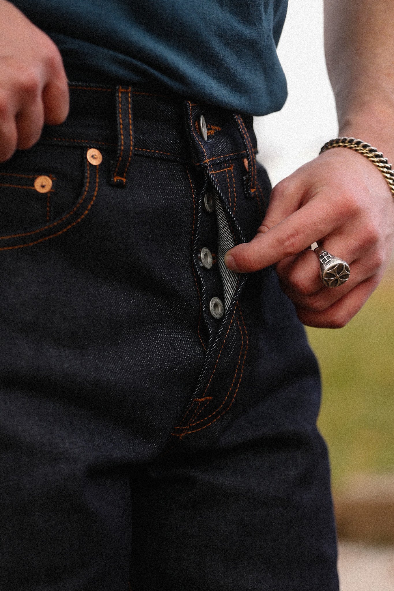 Close-up of person wearing dark indigo button-fly jeans with contrasting orange stitching and silver ring