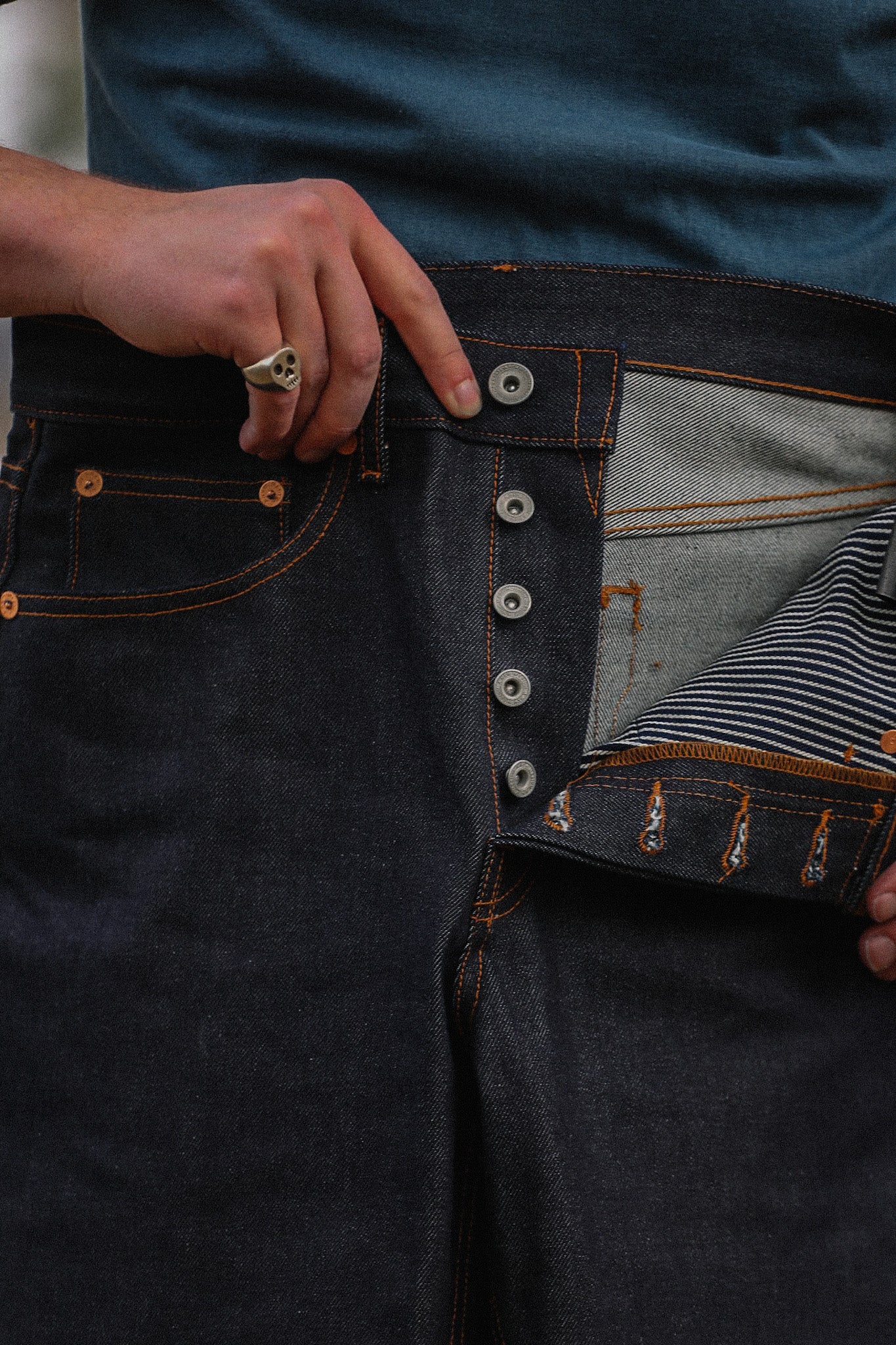 Close-up of hands showing button fly and interior striped lining of dark indigo wide straight jeans with orange stitching