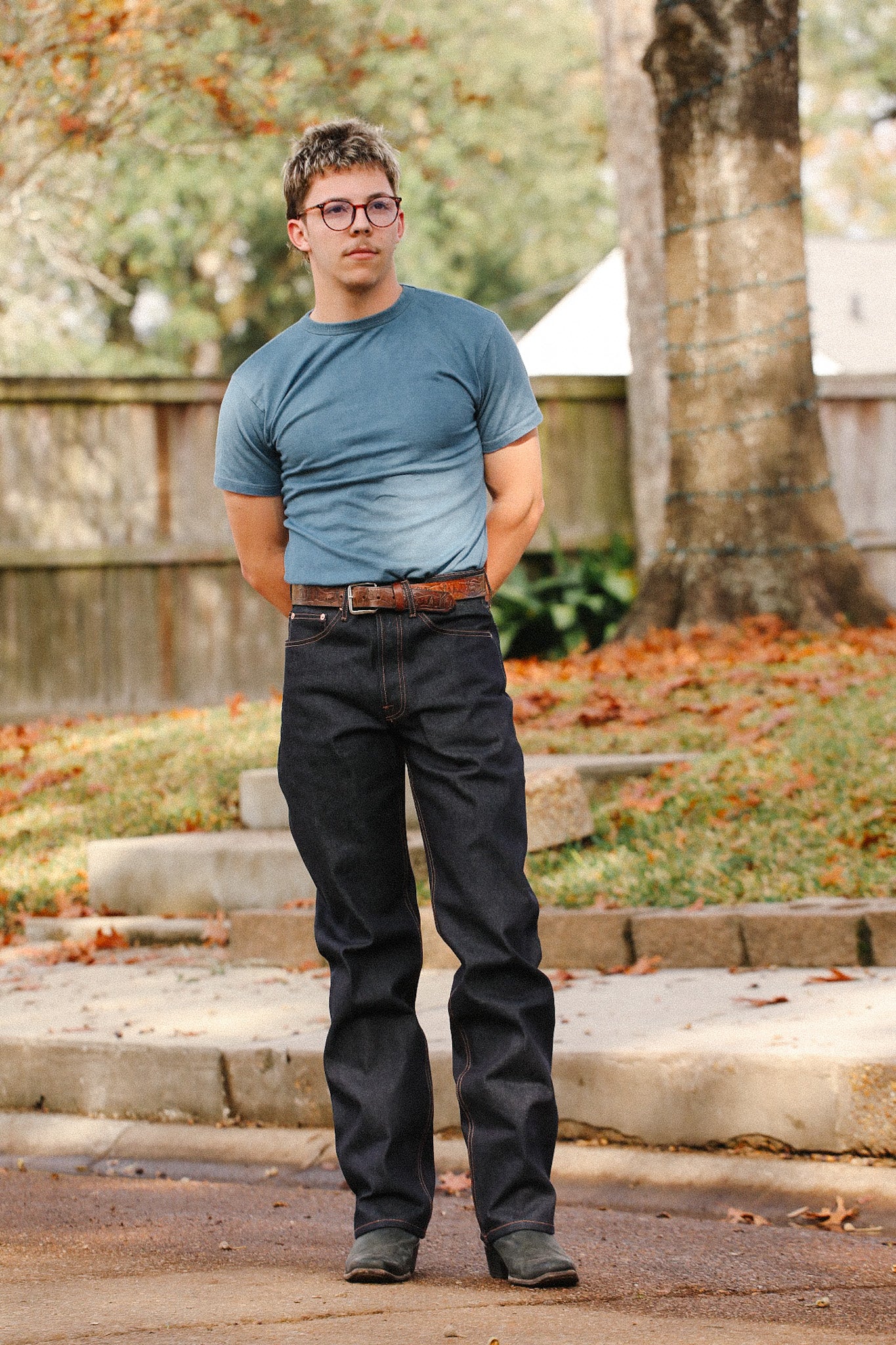 Man wearing wide straight 16.5oz indigo denim jeans with brown belt, blue t-shirt, and glasses outdoors