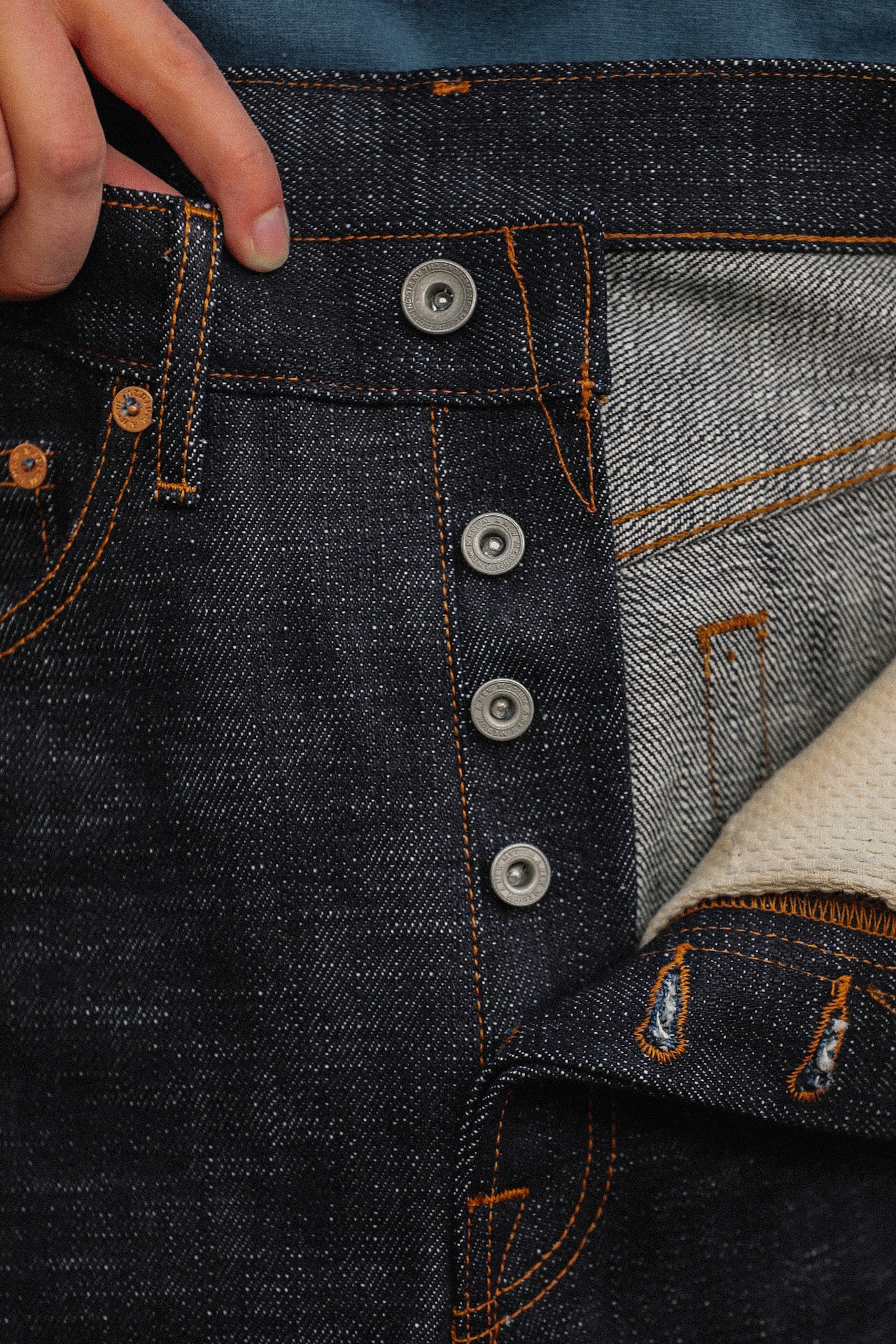 Close-up of hand holding dark indigo raw denim jeans with orange stitching and metal button fly