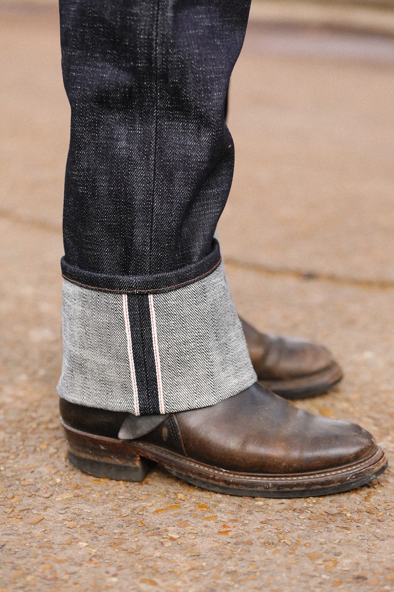 Close-up of folded dark indigo wide straight jeans cuff showing selvedge detail worn with brown leather boots on textured ground