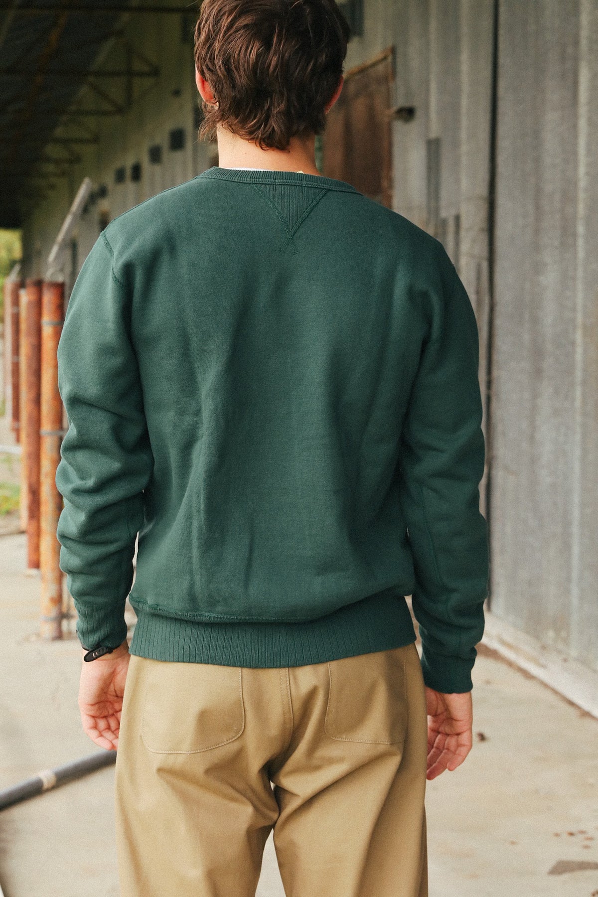 A man viewed from behind wearing a dark green crewneck sweatshirt and tan pants, standing in an industrial corridor with a gray metal wall.