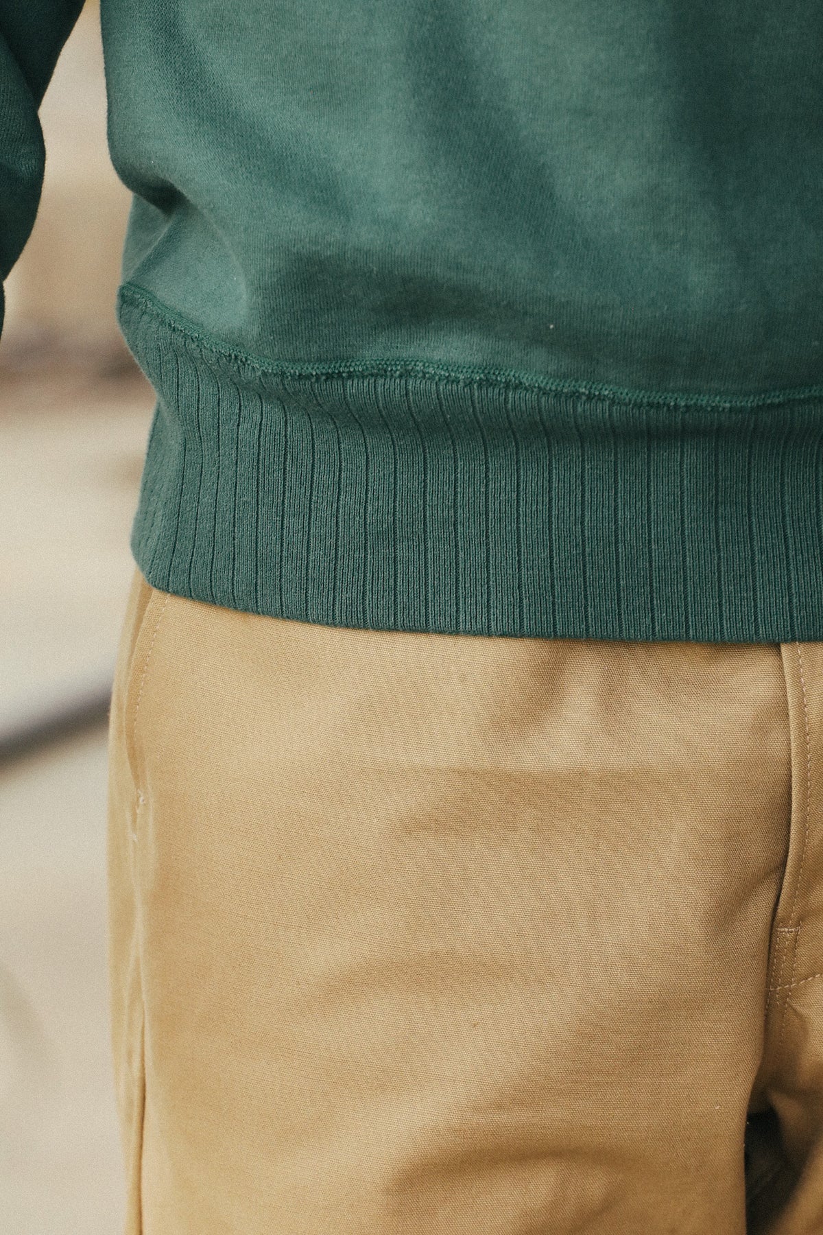 Close-up of a person wearing a dark teal-green ribbed knit sweater and tan pants, focusing on the hem of the sweater.