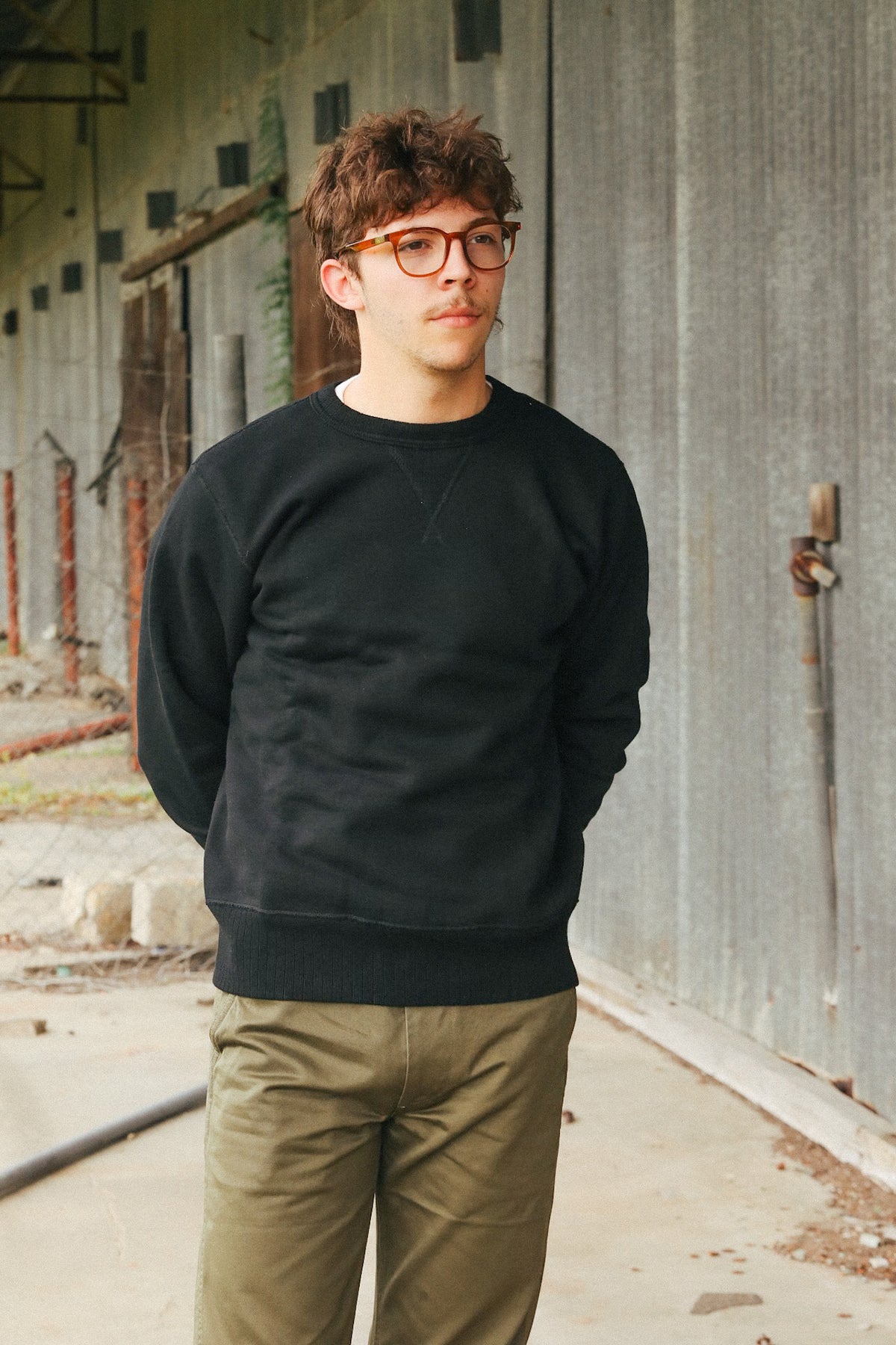 A man wearing a black crewneck sweatshirt and olive green pants, standing in front of a weathered gray metal wall.