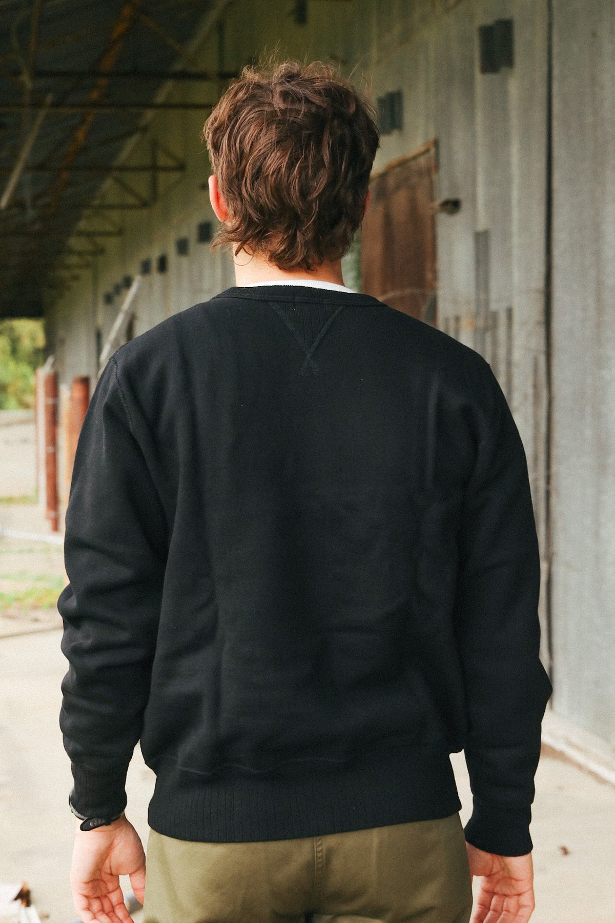 A man viewed from behind wearing a black crewneck sweatshirt and olive green pants, standing in an industrial corridor with a concrete wall.