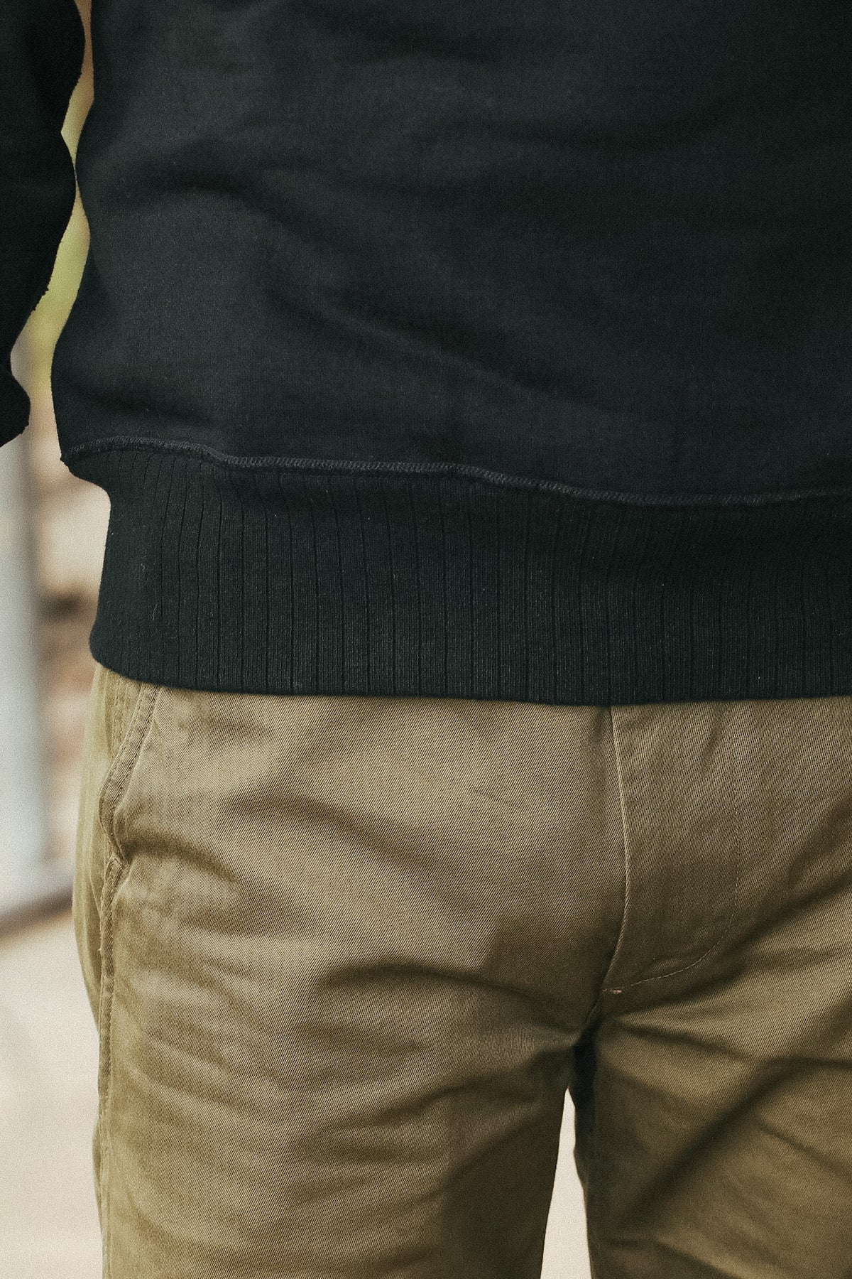 Close-up of a person wearing a black ribbed-knit crewneck sweatshirt and olive green pants.