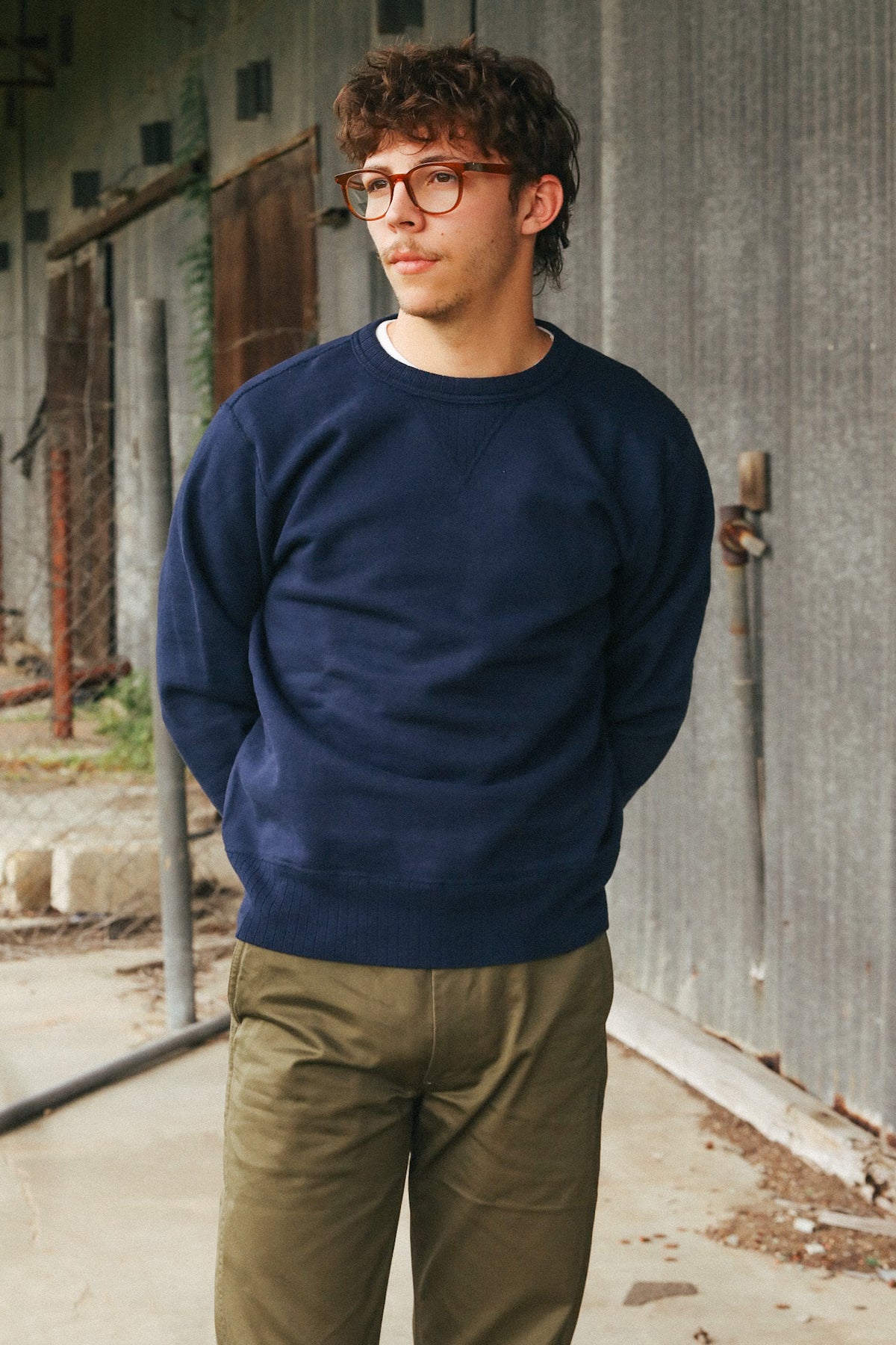 A man wearing a navy blue crewneck sweatshirt and olive green pants, standing outdoors against a gray corrugated metal wall.