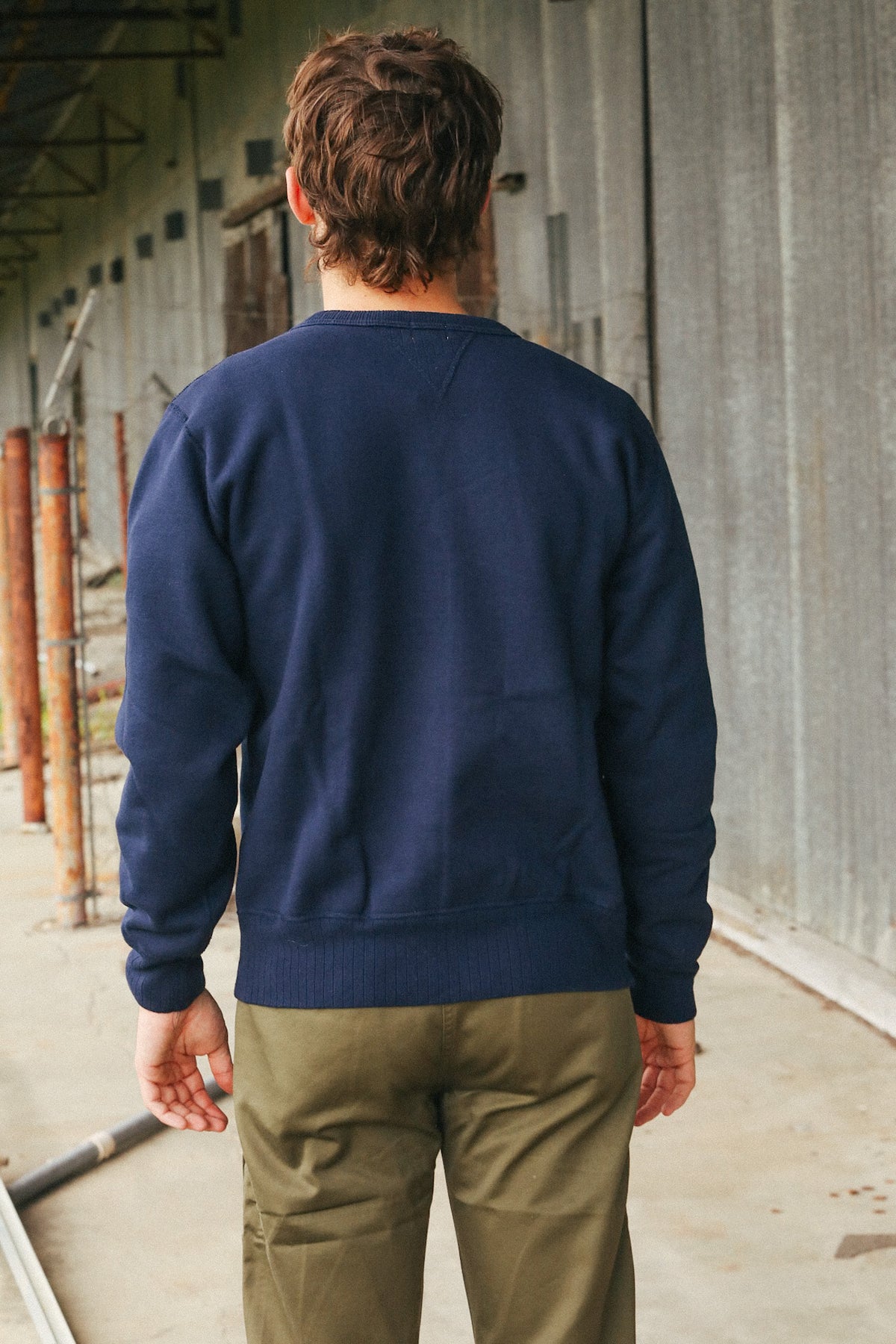 A man viewed from behind wearing a navy blue crewneck sweatshirt and olive green pants, standing in an industrial corridor with a corrugated metal wall.