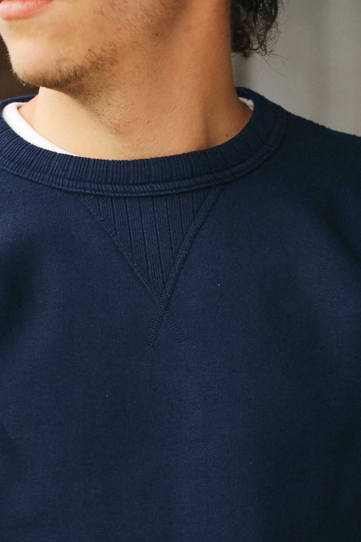 Close-up of a person wearing a navy crewneck sweatshirt over a white undershirt, showing the ribbed knit collar and chest detail.