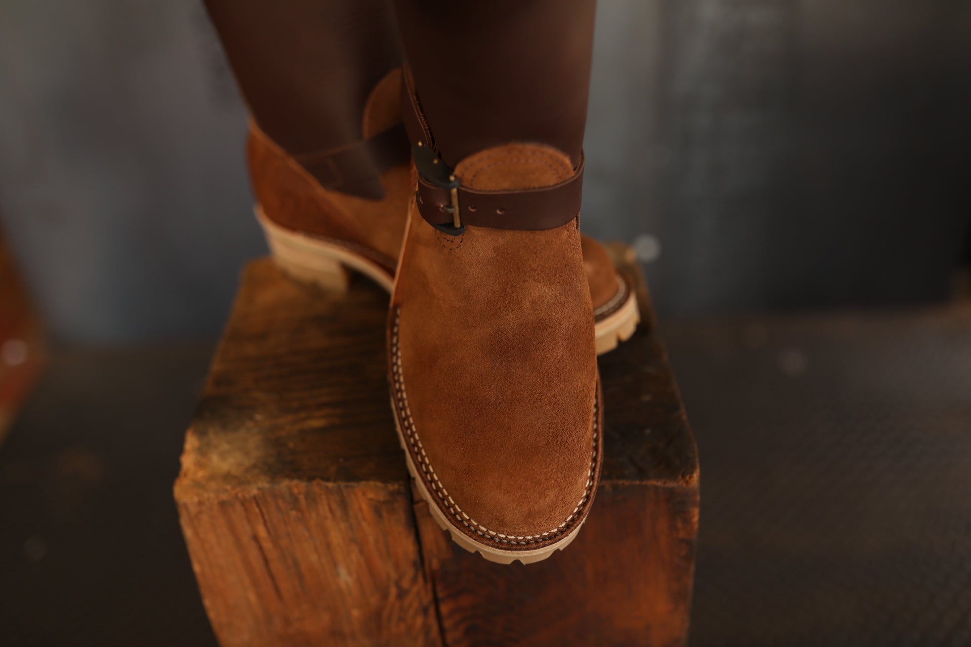 Brown suede engineer boots with leather strap and buckle on wooden block, industrial background