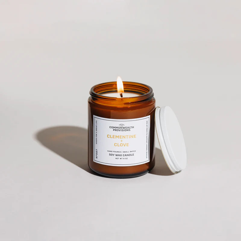 Lit amber glass soy wax candle labeled Clementine Clove by Commonwealth Provisions with white lid beside it on white surface