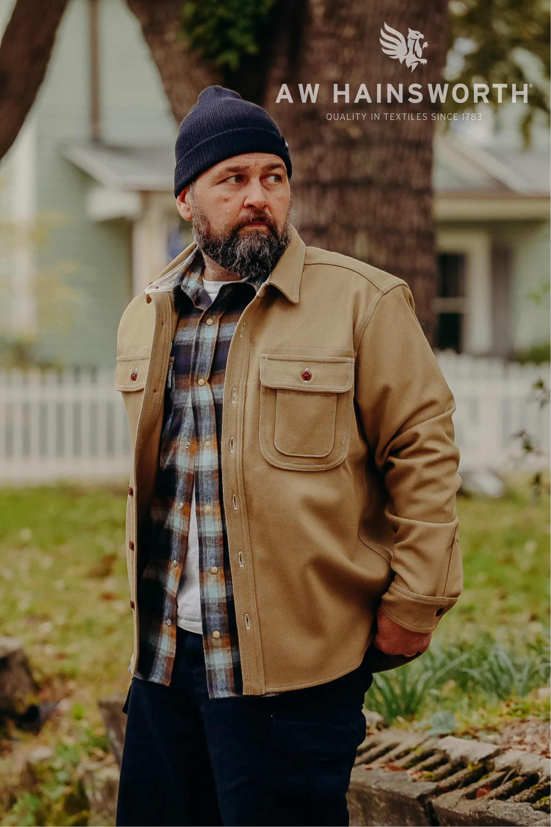 Man wearing AW Hainsworth tan work jacket and plaid shirt outdoors near house and tree