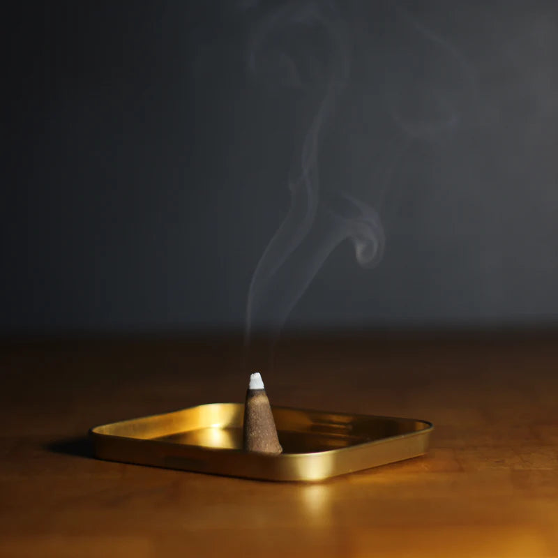 Burning incense cone emitting smoke on a gold metal tray on wooden surface