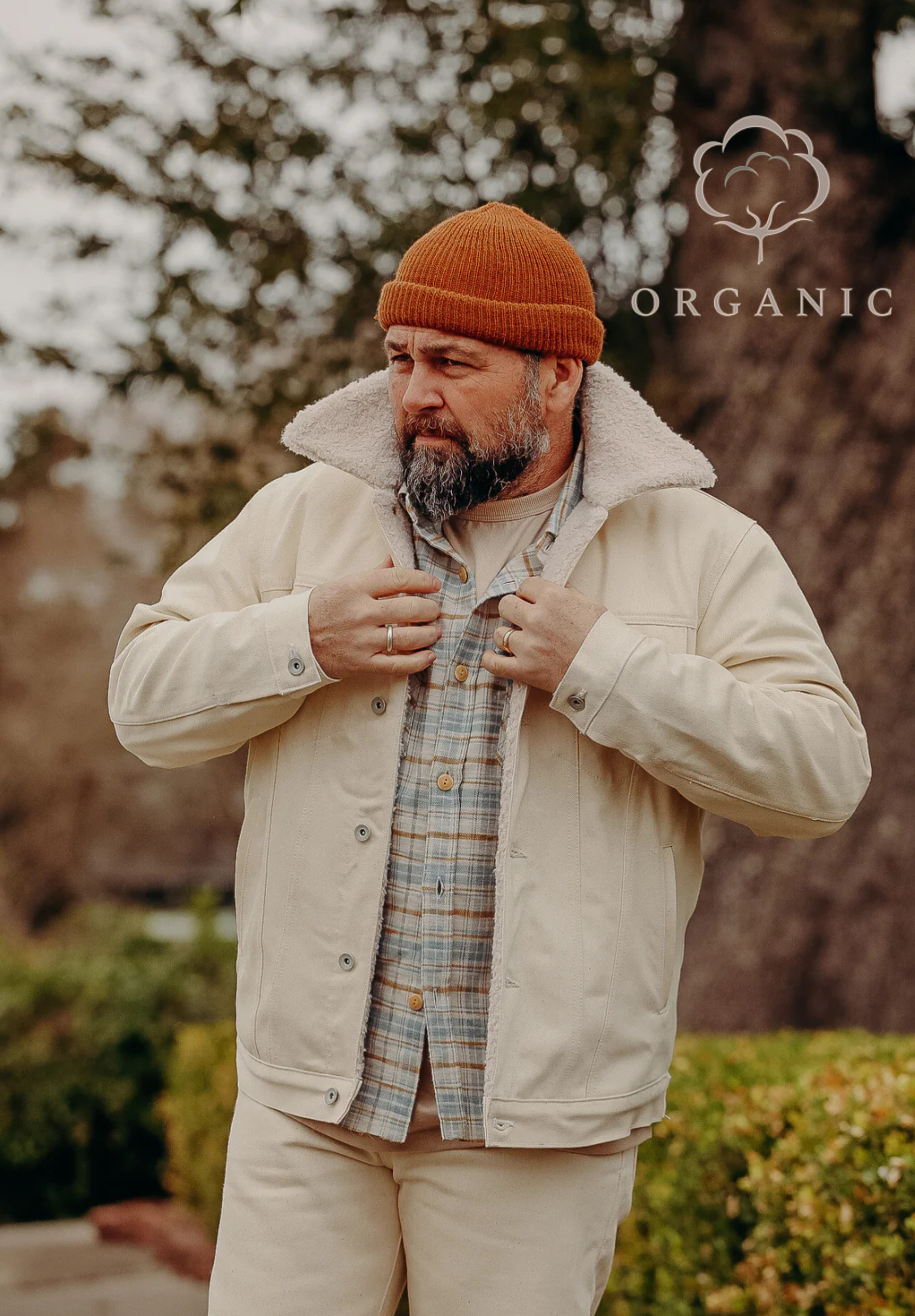 Bearded man wearing beige sherpa jacket, plaid shirt, and orange beanie outdoors with organic cotton logo