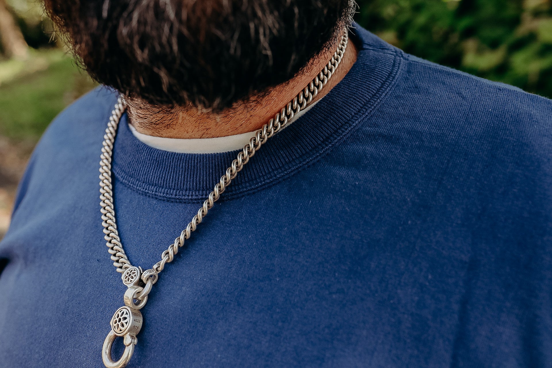 Close-up of a man wearing a silver chain necklace with detailed clasps over a blue crewneck shirt