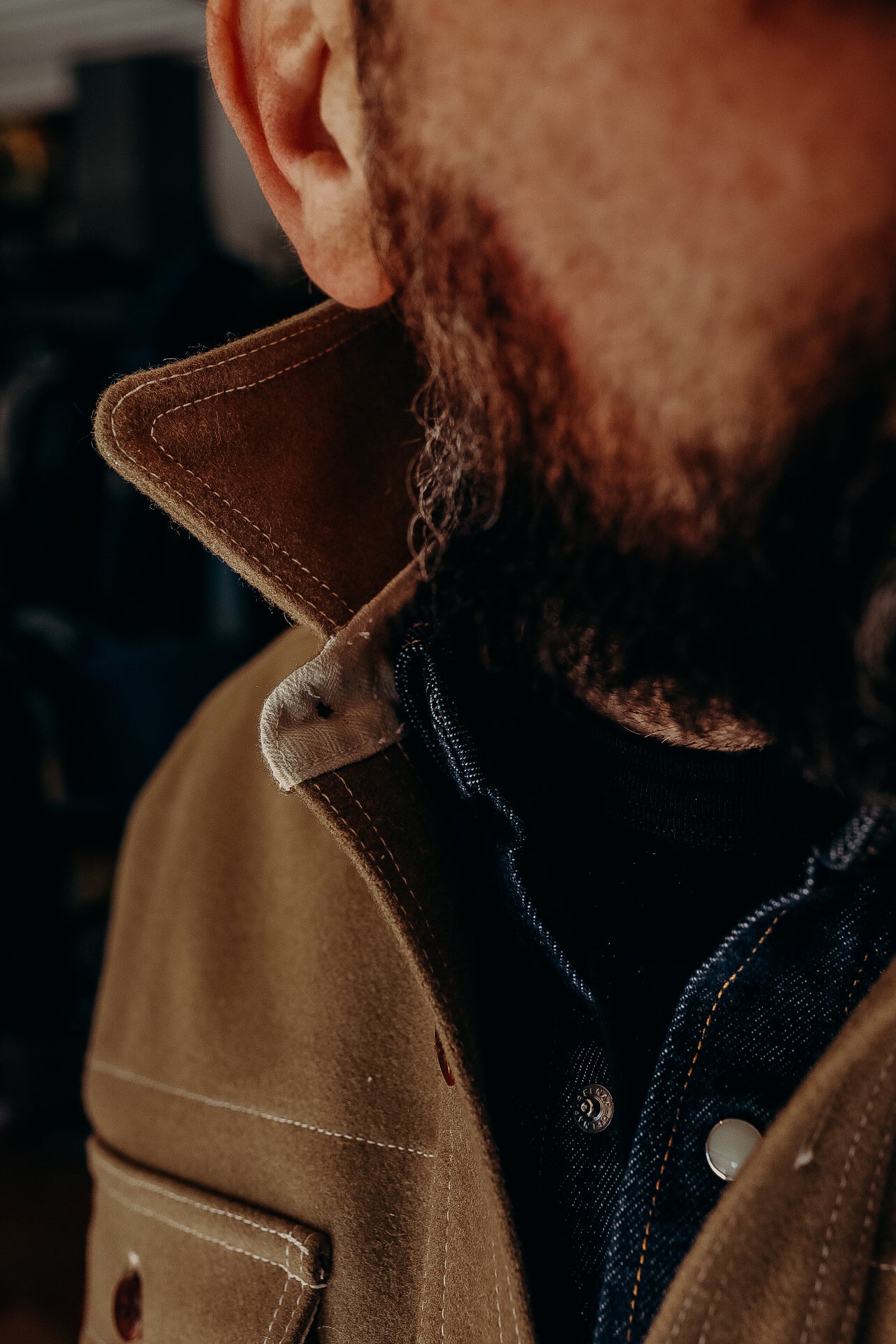 Close-up of man wearing tan work jacket with raised collar and denim shirt, showing beard details