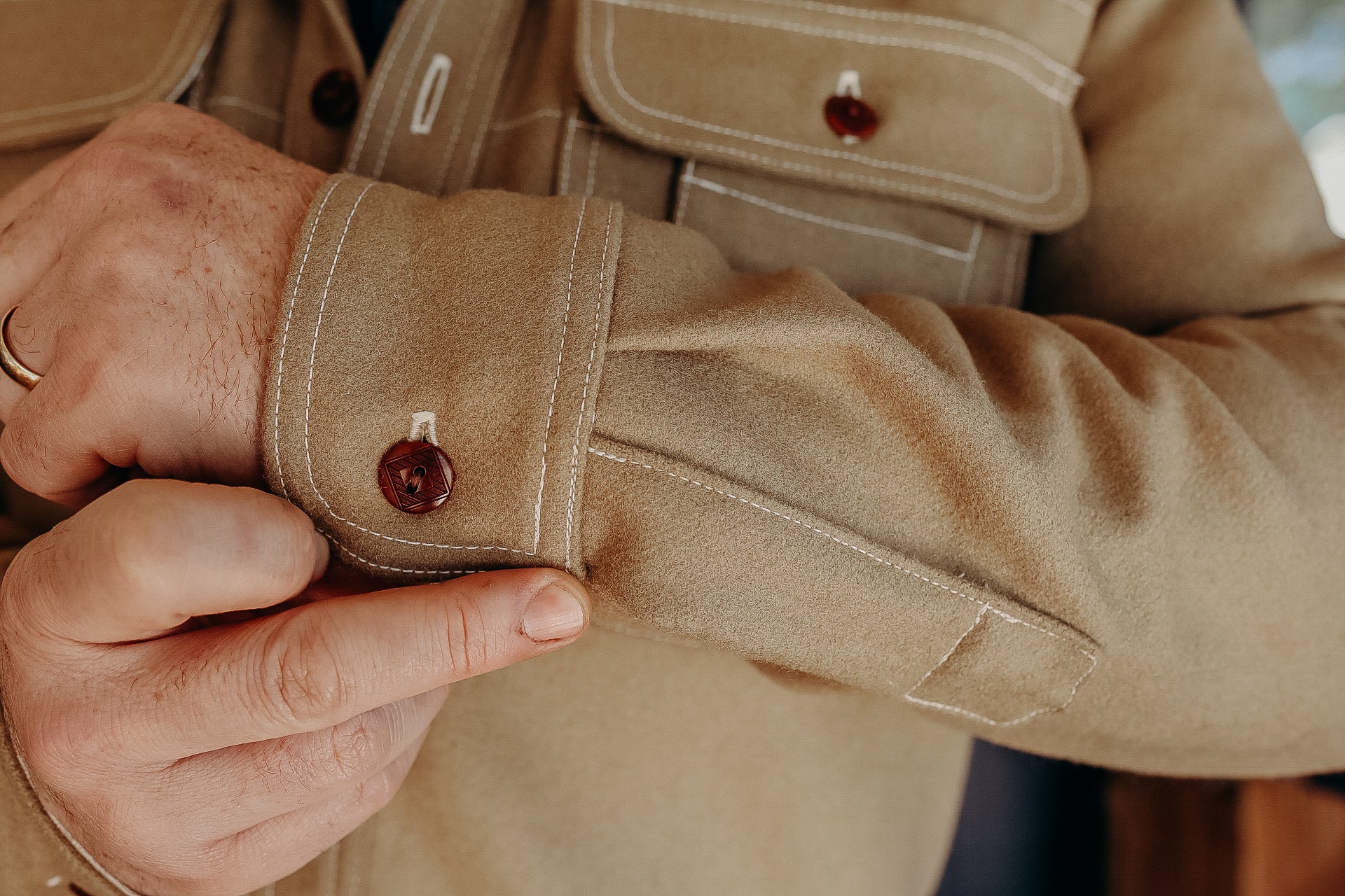 Close-up of a person buttoning the cuff of a beige suede jacket with brown buttons and white stitching