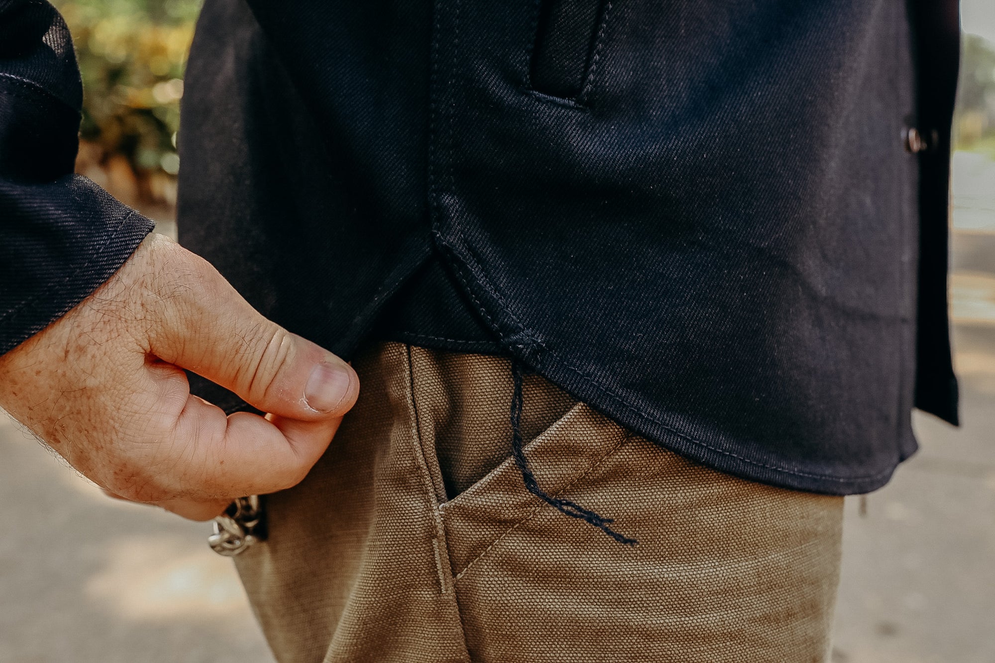 Close-up of a hand holding a black work shirt hem over brown canvas pants pocket outdoors