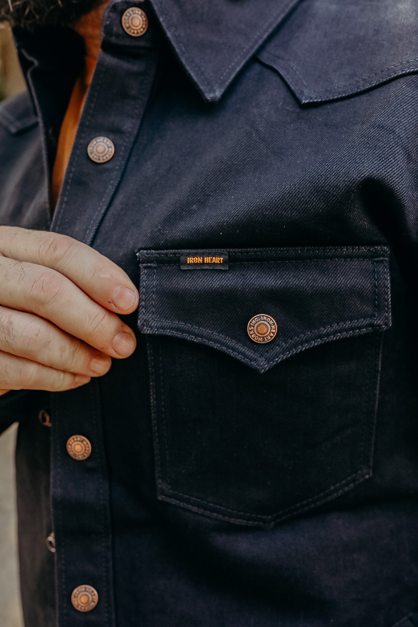 Close-up of a hand holding the chest pocket flange of a dark blue Iron Heart denim jacket with branded copper buttons