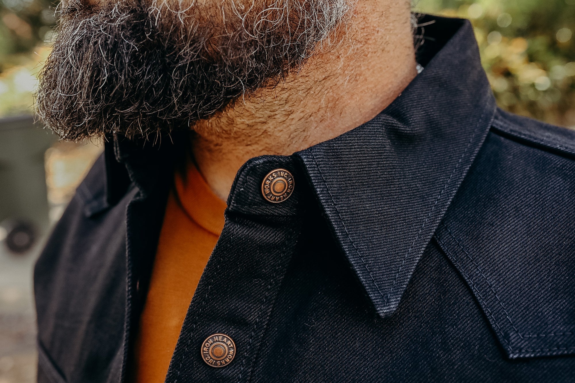 Close-up of a man with a salt-and-pepper beard wearing a black denim jacket with branded metal buttons and an orange shirt