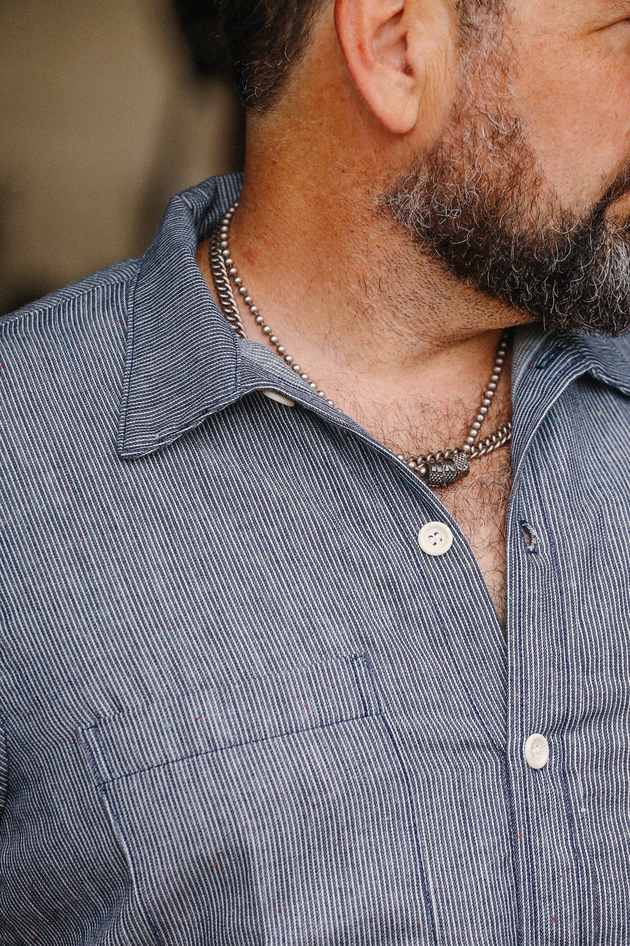 Close-up of a man wearing a blue striped v-short sleeve shop shirt with white buttons and a silver chain necklace