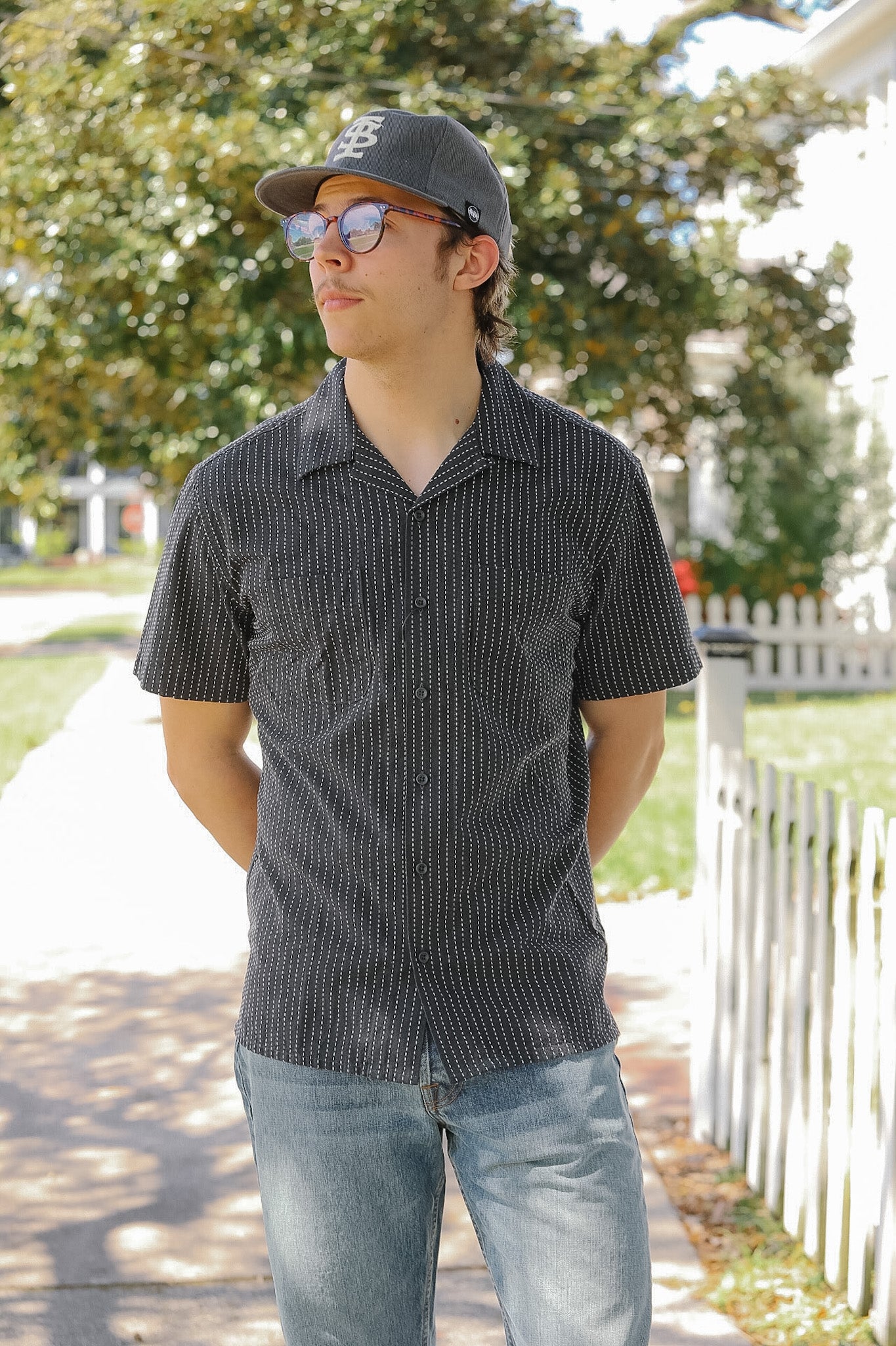 Man wearing a dark patterned shirt and cap outdoors with trees and a white fence in the background