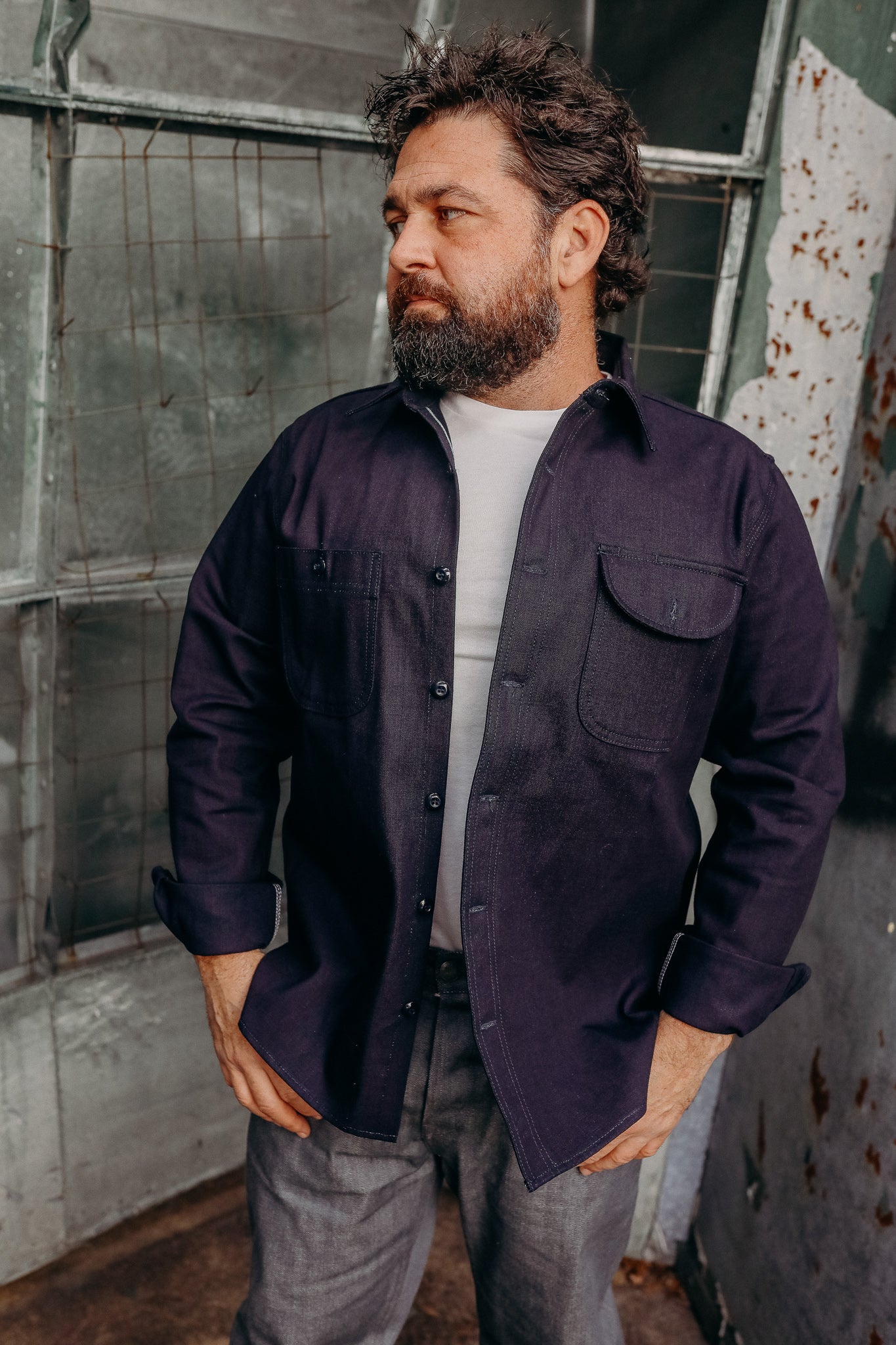 Bearded man in navy work shirt and white tee posing in rustic industrial setting with metal doors