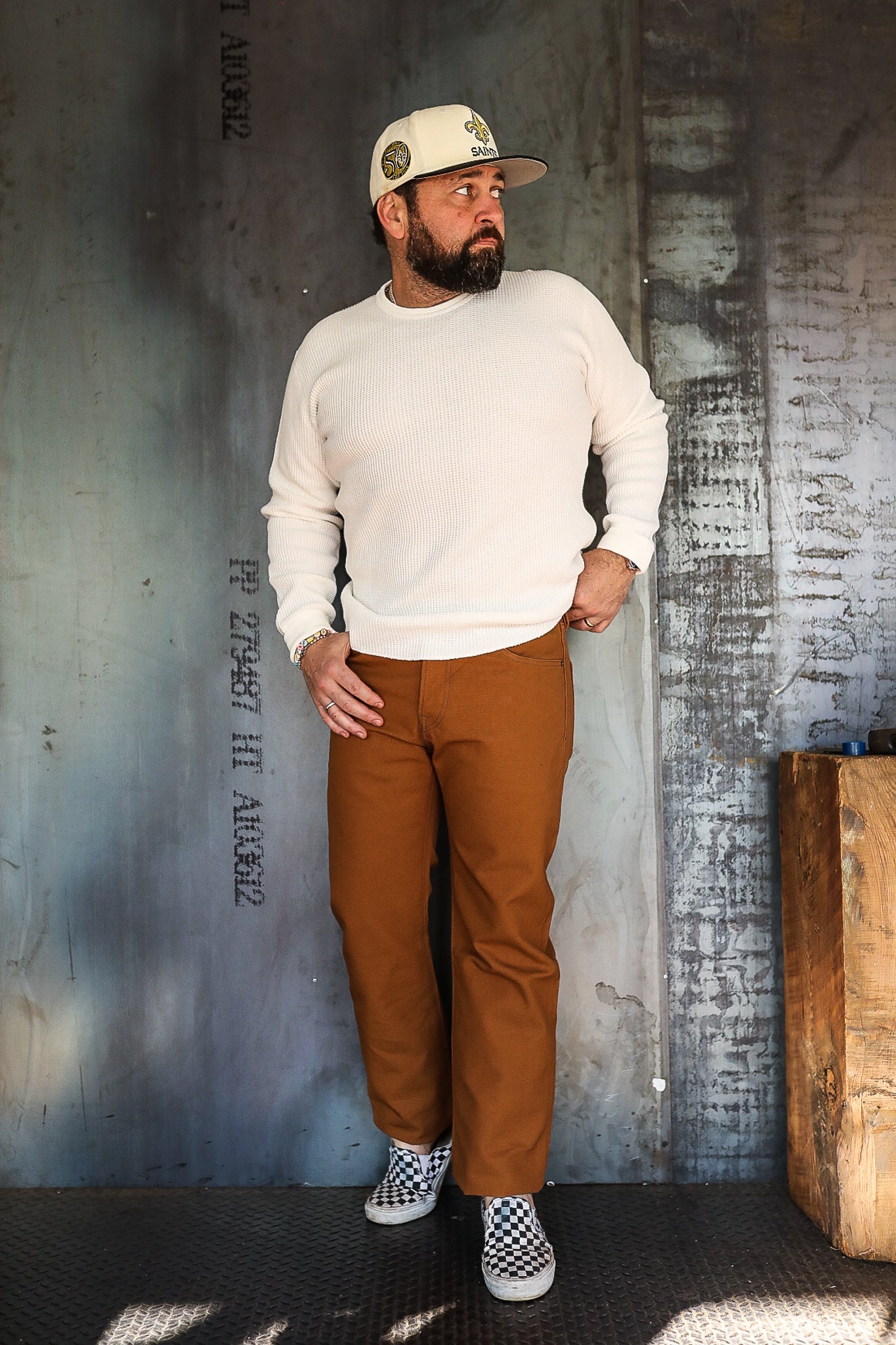 Man wearing white knit sweater, brown work pants, and checkered slip-on shoes in industrial setting