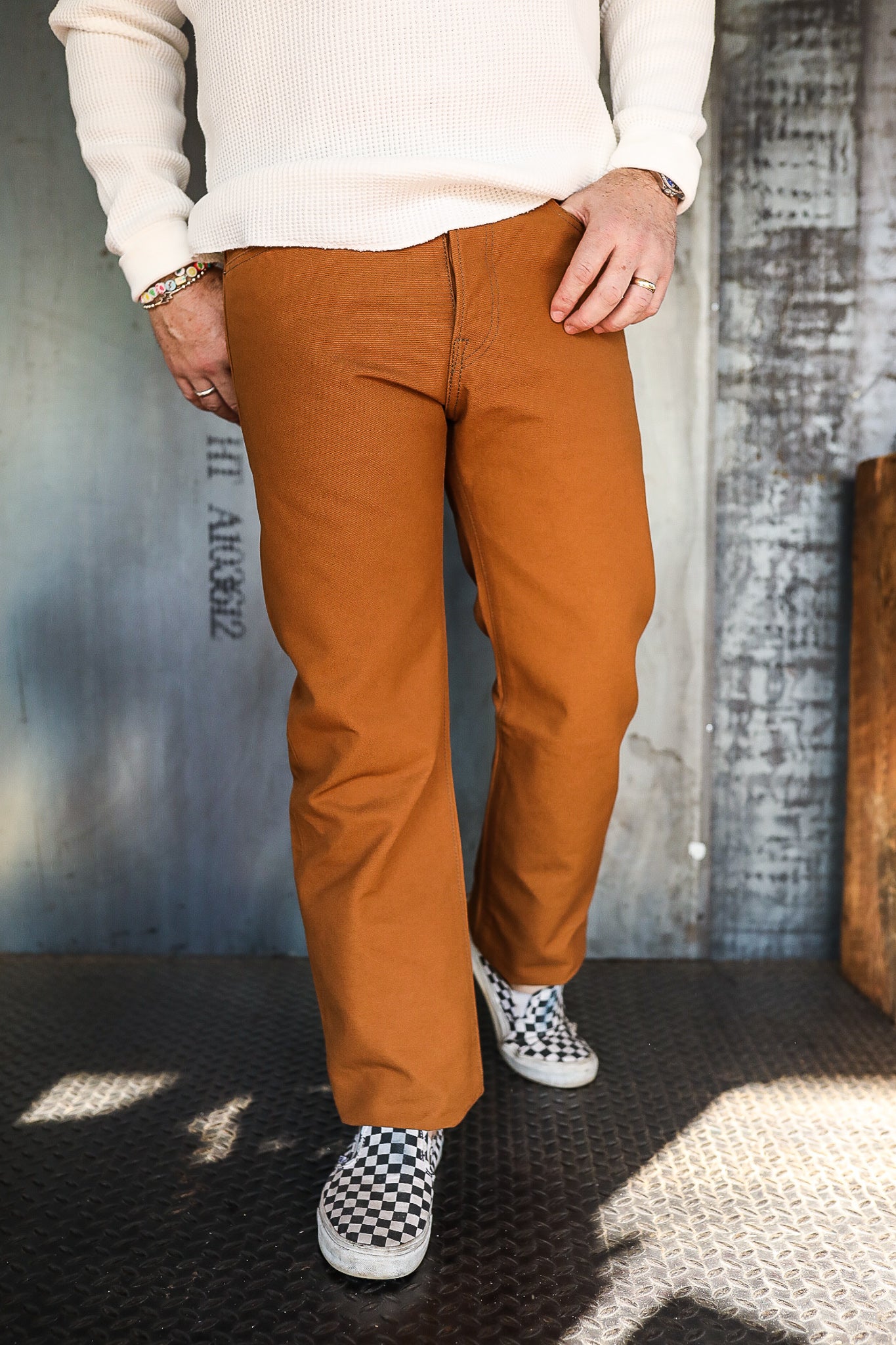 Man wearing rust-colored casual pants with a white waffle knit sweater and black-and-white checkerboard slip-on shoes on industrial floor