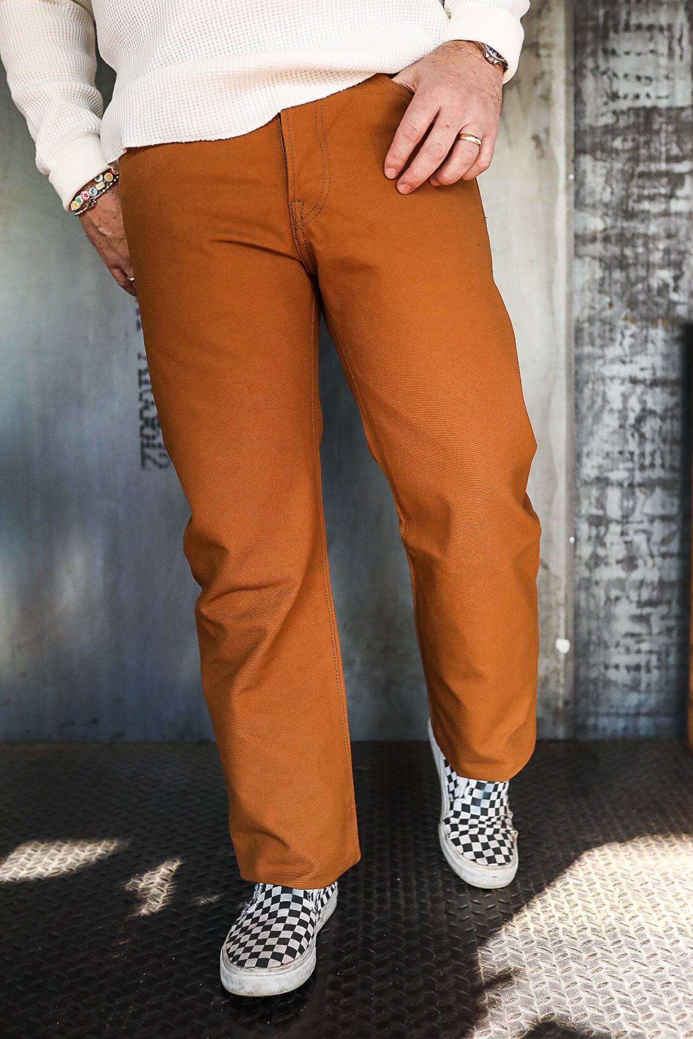 Man wearing brown ironwork pants with black and white checkered slip-on shoes standing on industrial metal floor