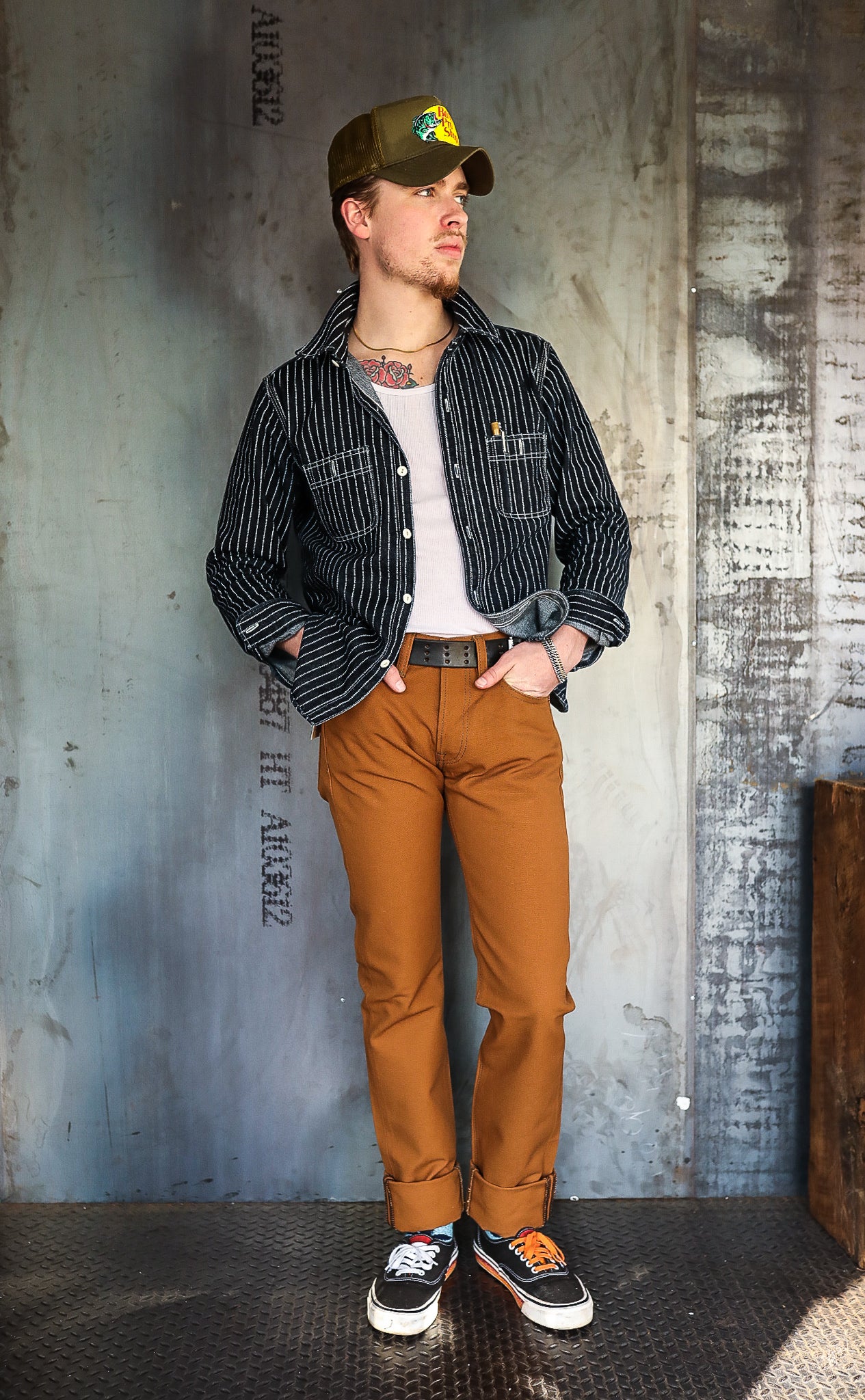 Man wearing striped black jacket, white tank top, brown pants with rolled cuffs, and black Vans sneakers